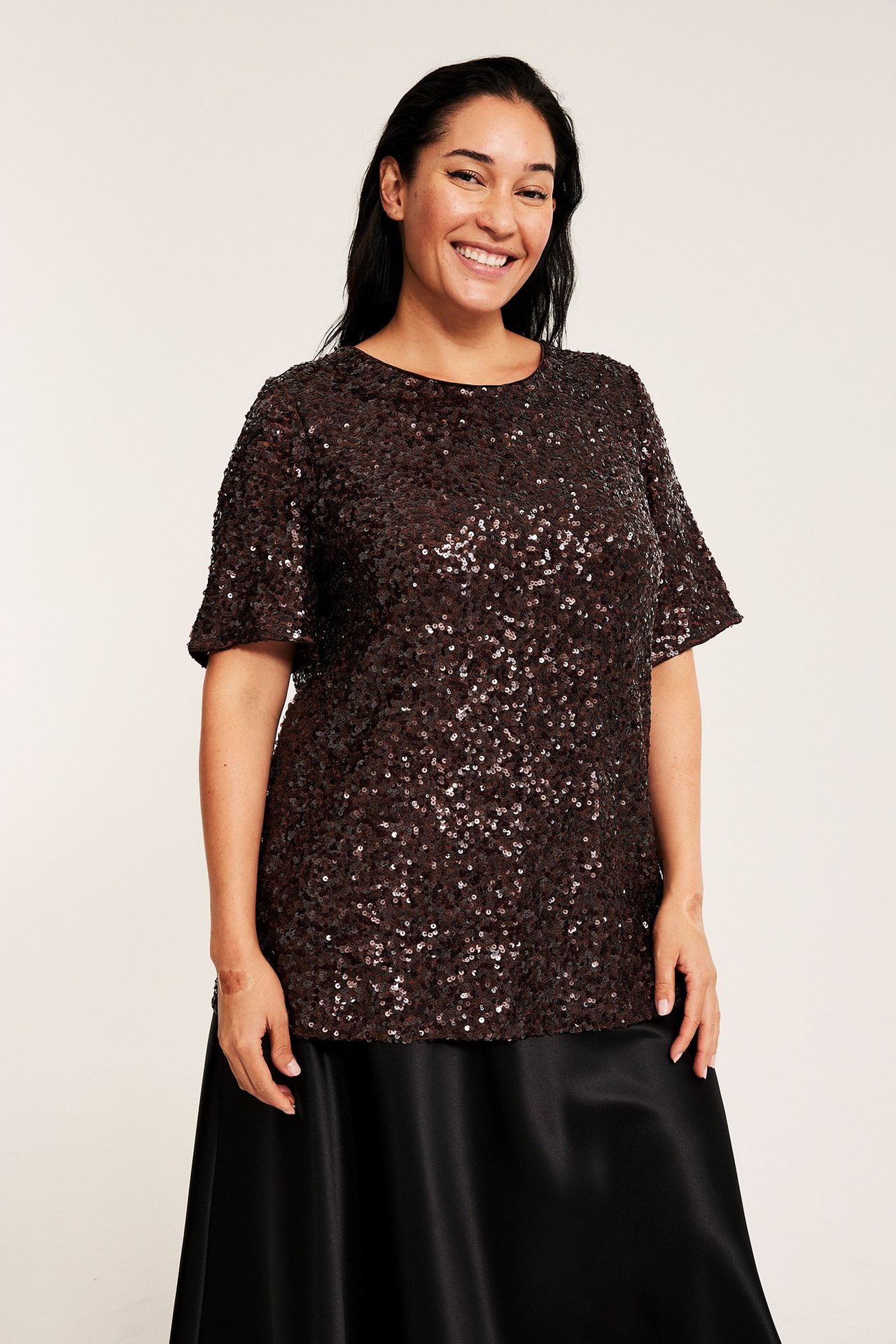 Edestä, hymyilevä nainen yllään ruskea paljettitoppi lyhyillä hihoilla ja pitkä musta hame.
