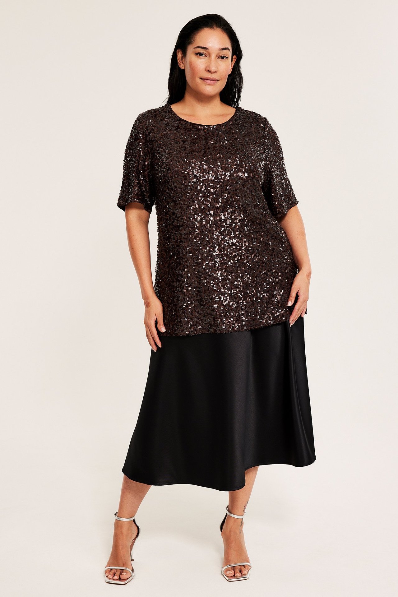 Edestä: Nainen ruskeassa paljettitopissa, mustassa midihameessa ja hopeisissa sandaaleissa.