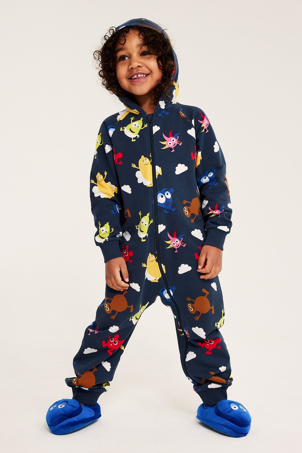Leende barn bär mörkblå mönstrad Babblarna-onesie med huva och blå tofflor.
