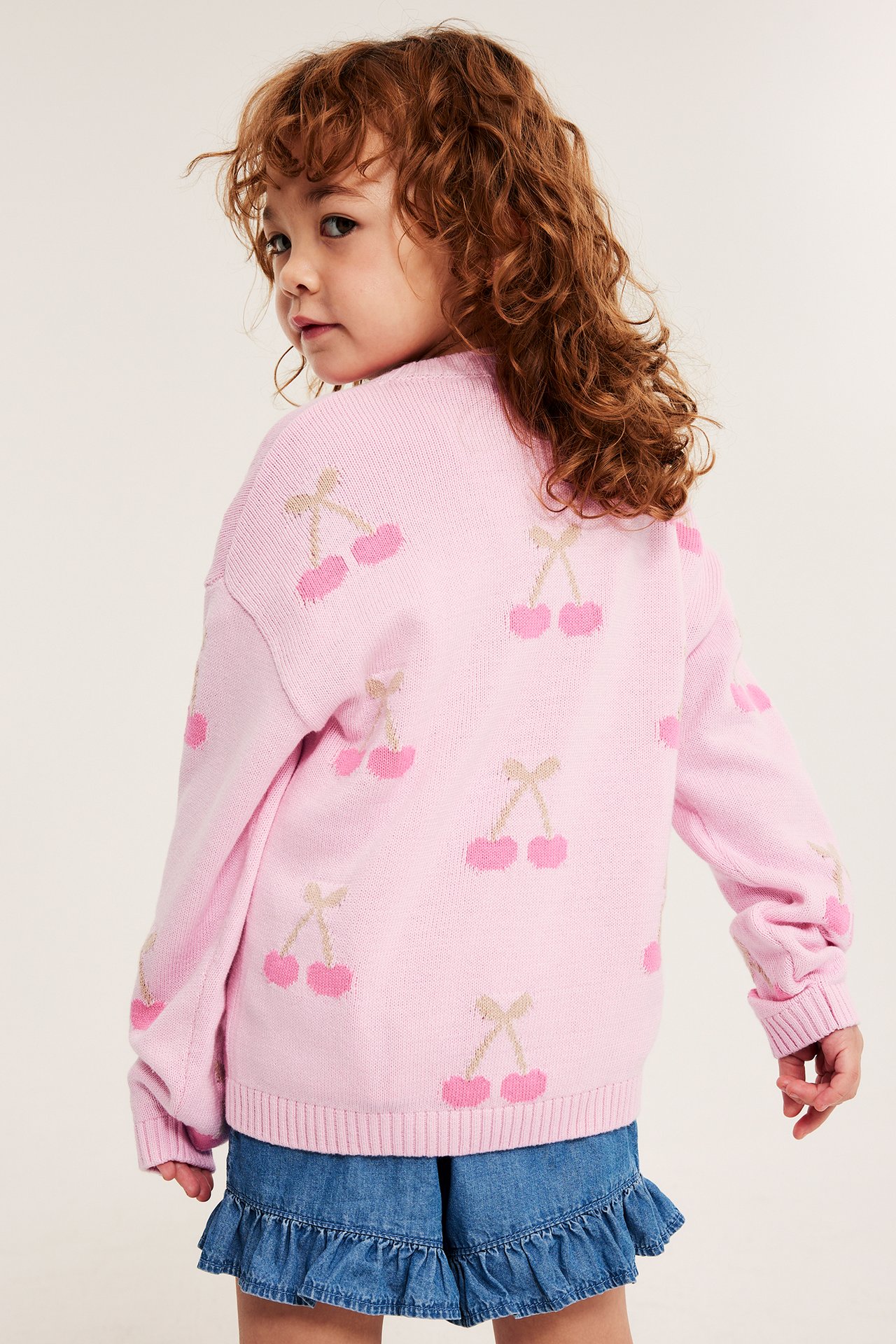 Ryggvy av ett barn i rosa mönstrad stickad bomullströja med körsbär och blå denim volangshorts.
