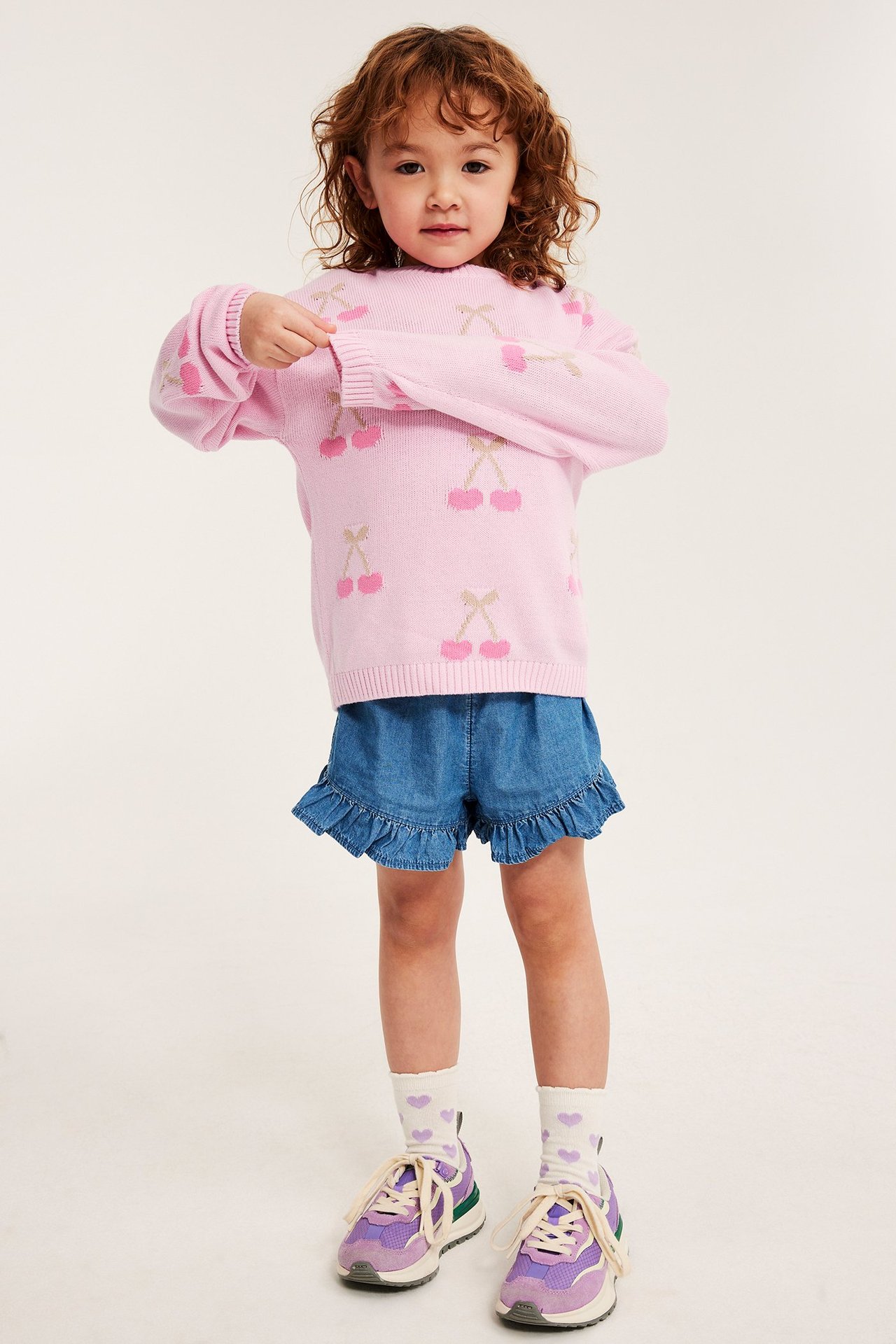 Framifrån, ett barn i rosa mönsterstickad bomullströja med körsbär, blå jeansshorts och lila sneakers.