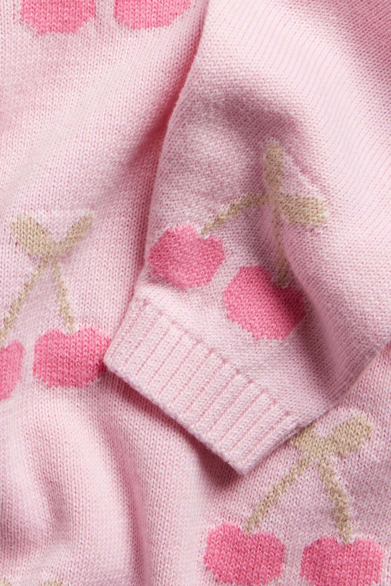 Lähikuva vaaleanpunaisesta puuvillaneuleesta tummanpunaisella ja beigellä kirsikkakuviolla ja resorihihansuulla.