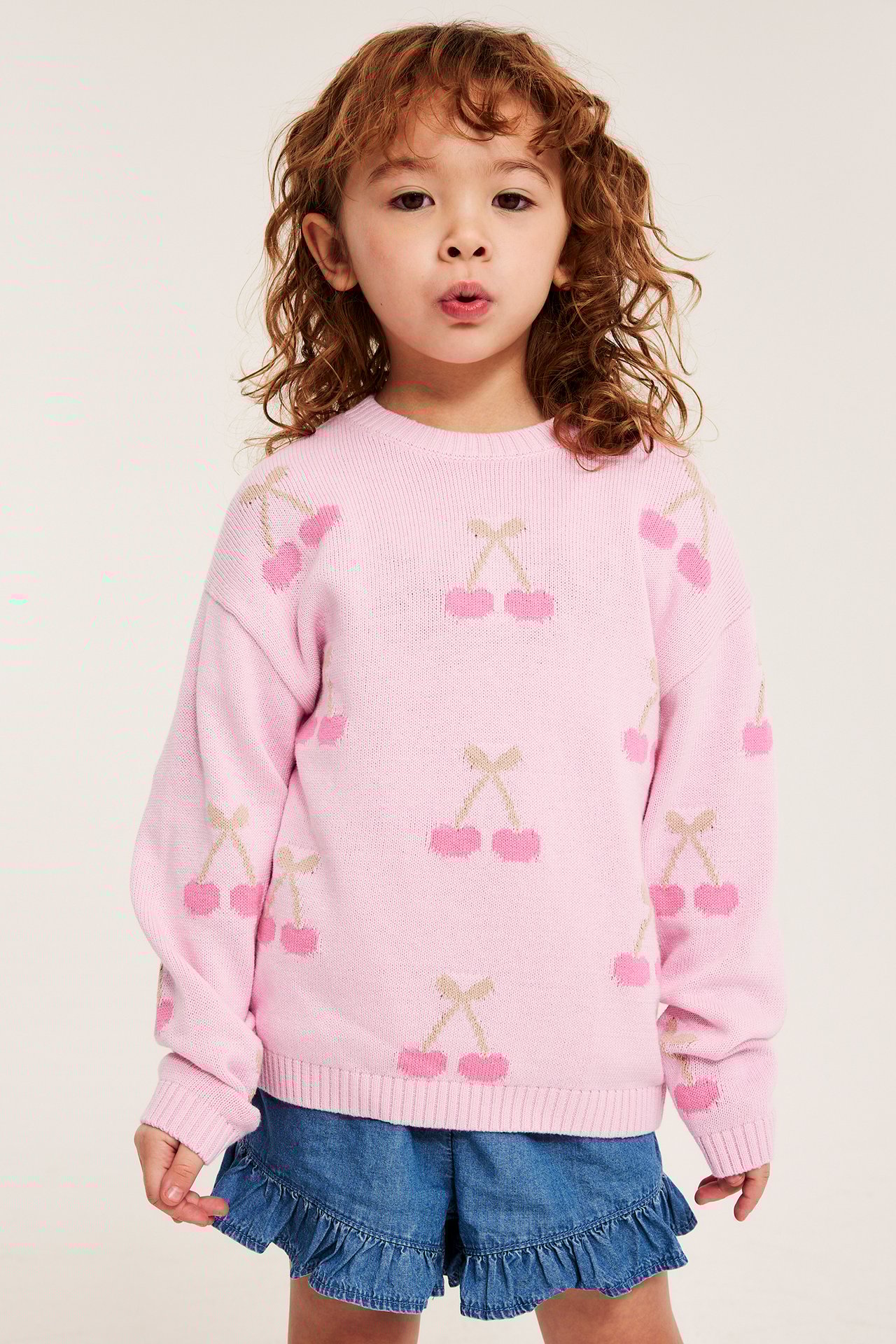 Frontvy av ett barn i en rosa mönsterstickad bomullströja med körsbär och blå jeansshorts.
