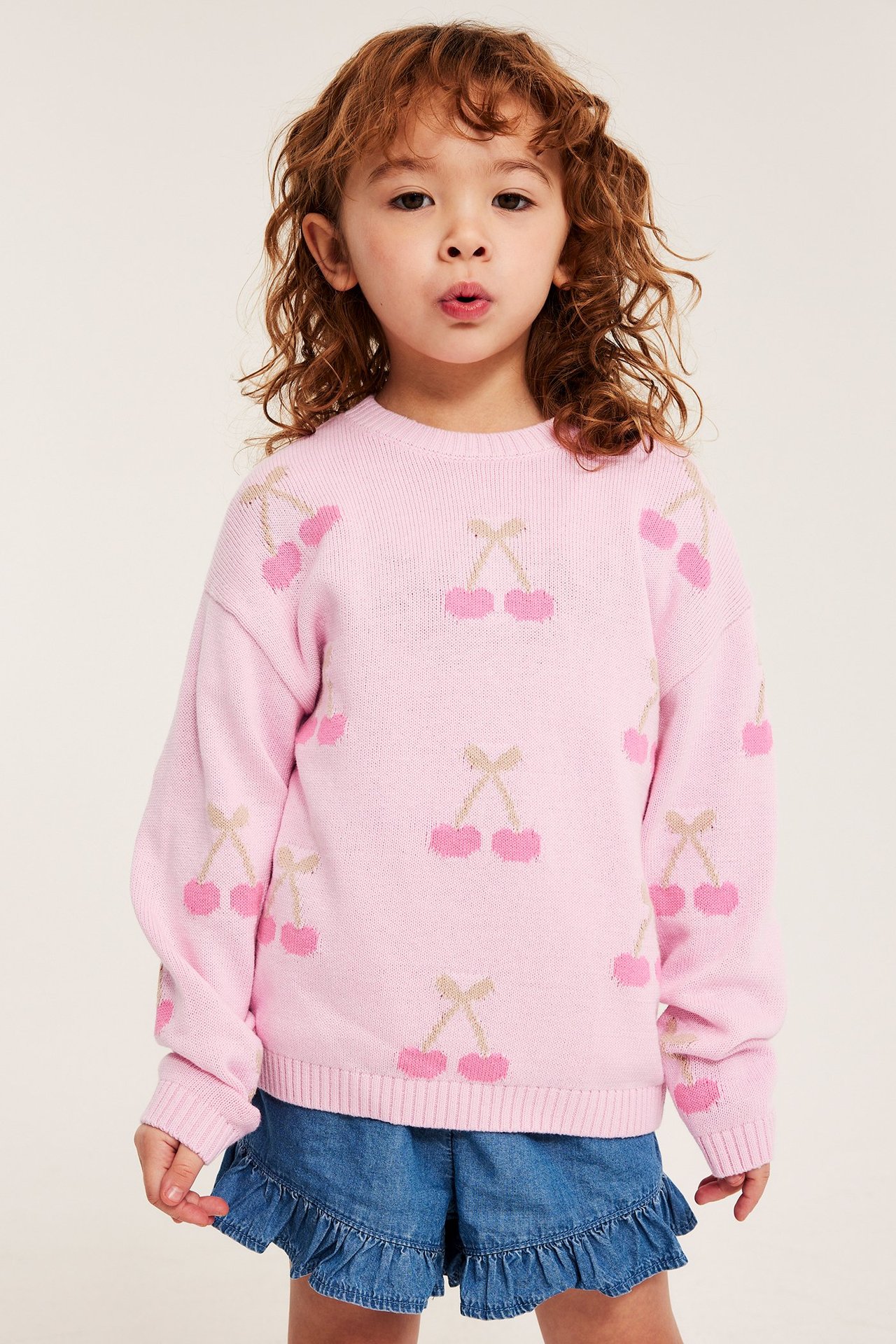 Forfra, et barn i en rosa mønsterstrikket bomullsgenser med kirsebær og blå dongerishorts.