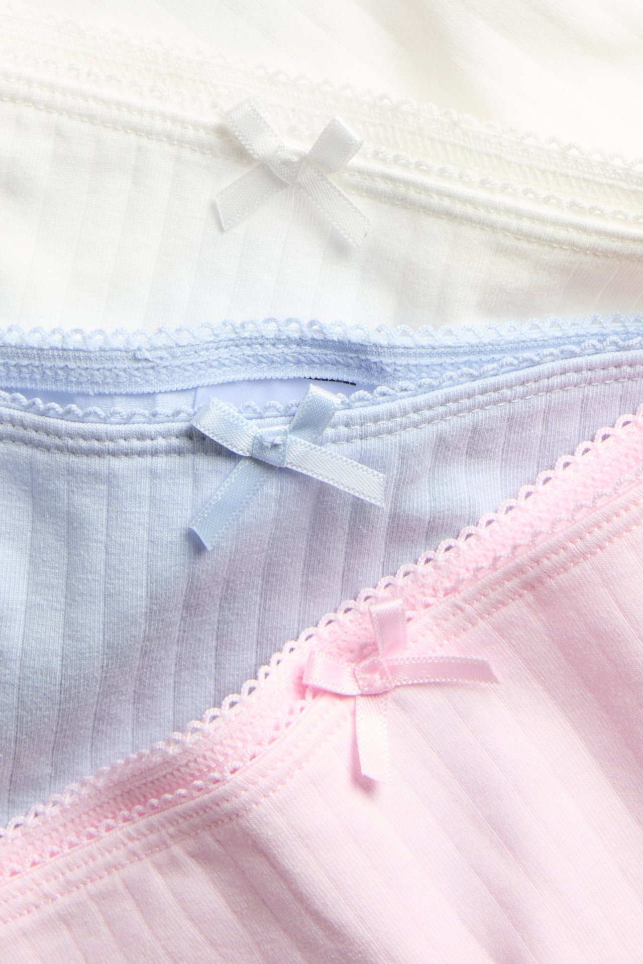 Zbliżenie trzech prążkowanych bokserek bawełnianych w kolorze różowym, jasnoniebieskim i białym, z lamówką picot i kokardkami.