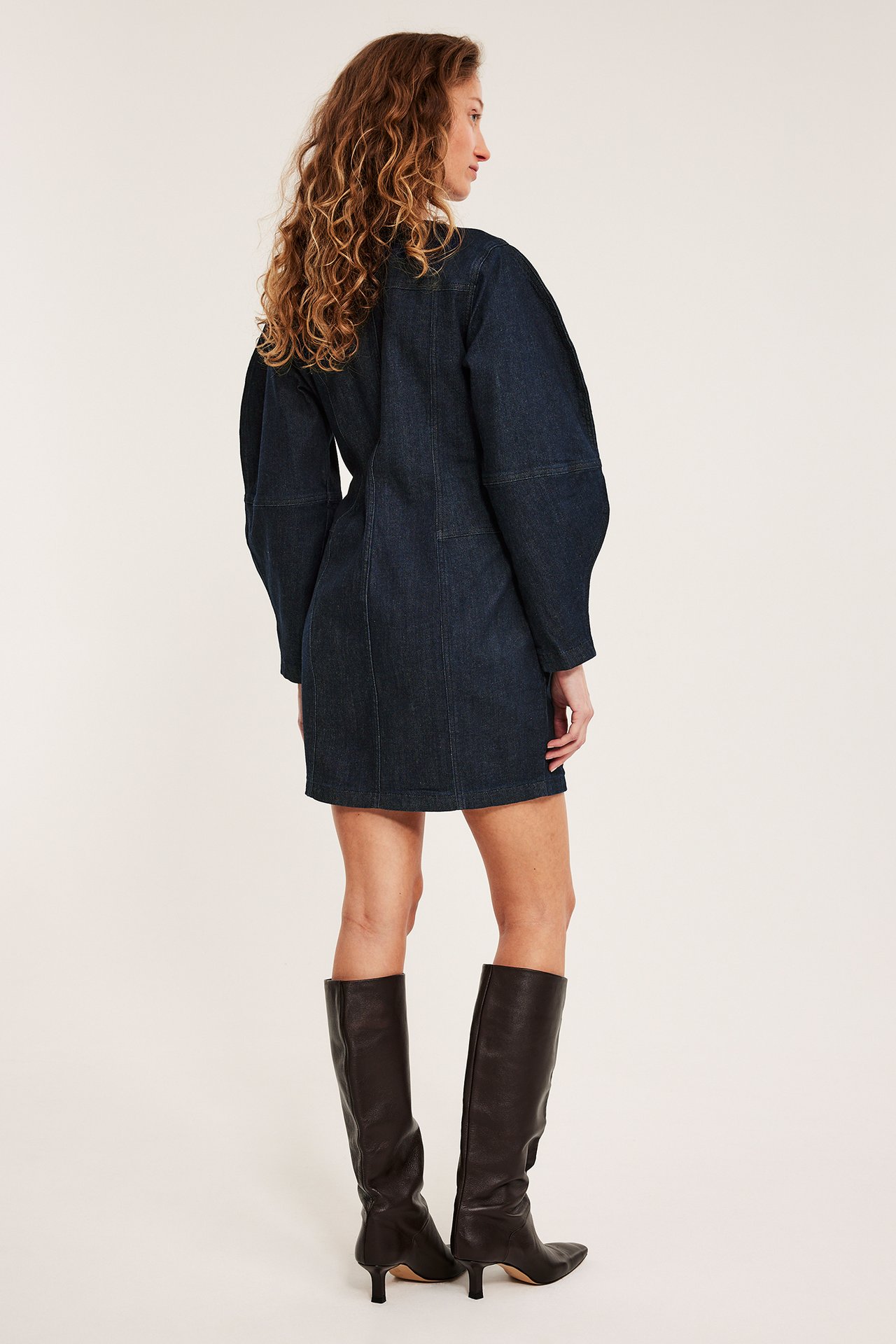 Bakfra: kvinne i en tettsittende mørk jeanskjole med voluminøse ermer og mørkebrune knestøvler.