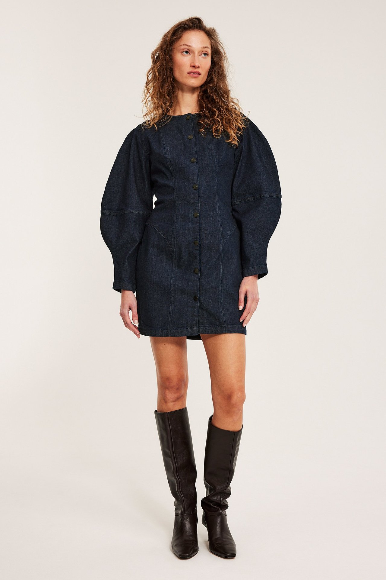 Frontutsikt: Kvinne i mørk, ettersittende jeanskjole med voluminøse ermer, mørke knapper og svarte knestøvler.