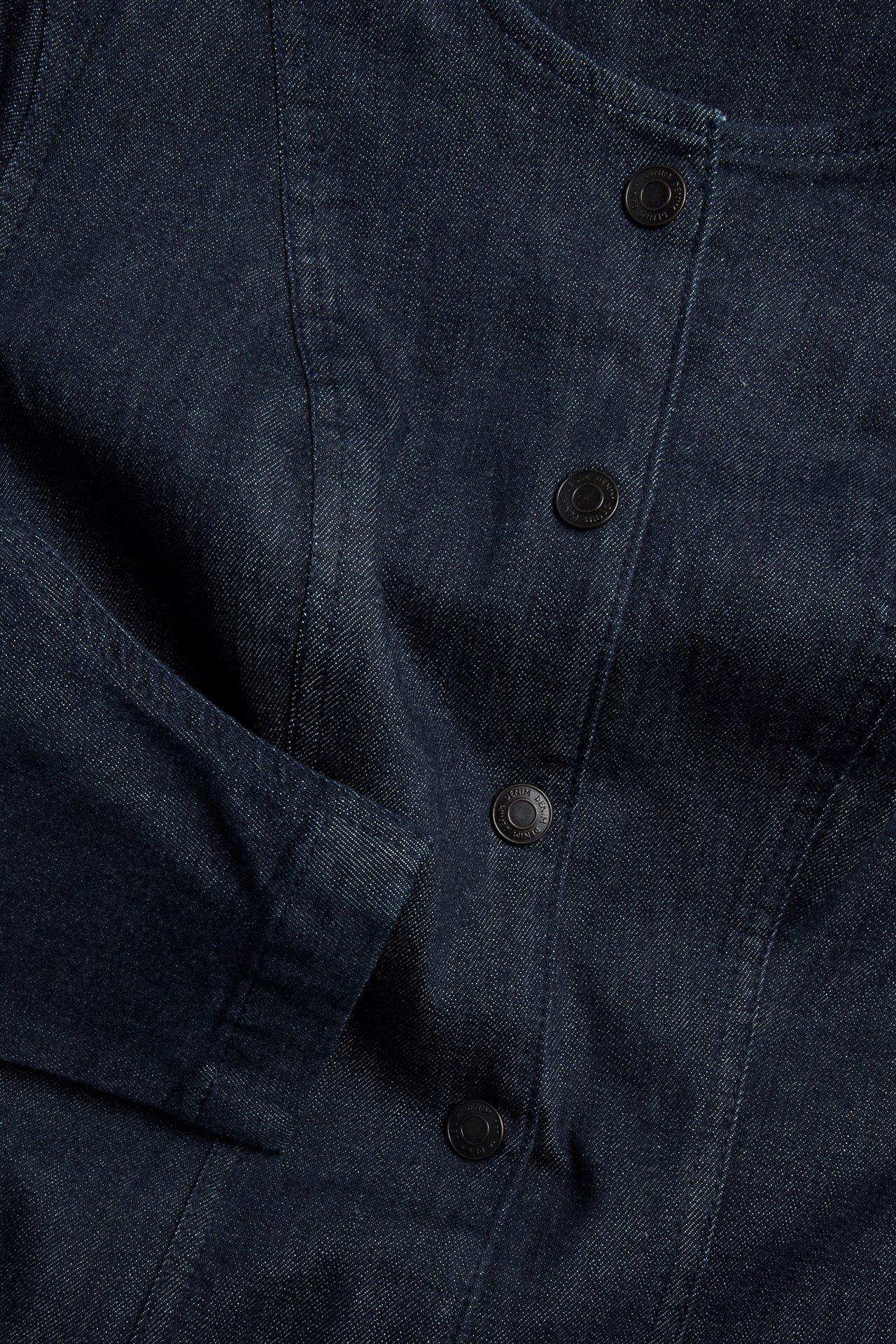 Nærbilde forfra av en mørk denimkjole med svarte knapper og skulpturert erme.