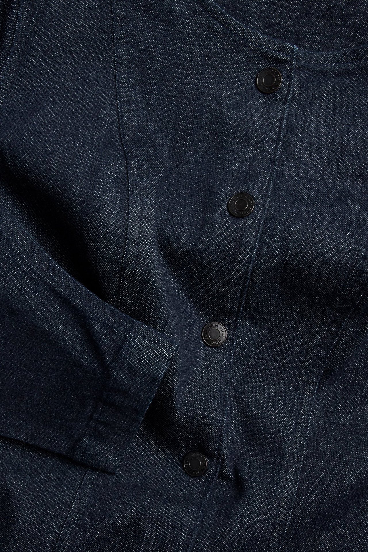 Nærbilde forfra av en mørk denimkjole med svarte knapper og skulpturert erme.