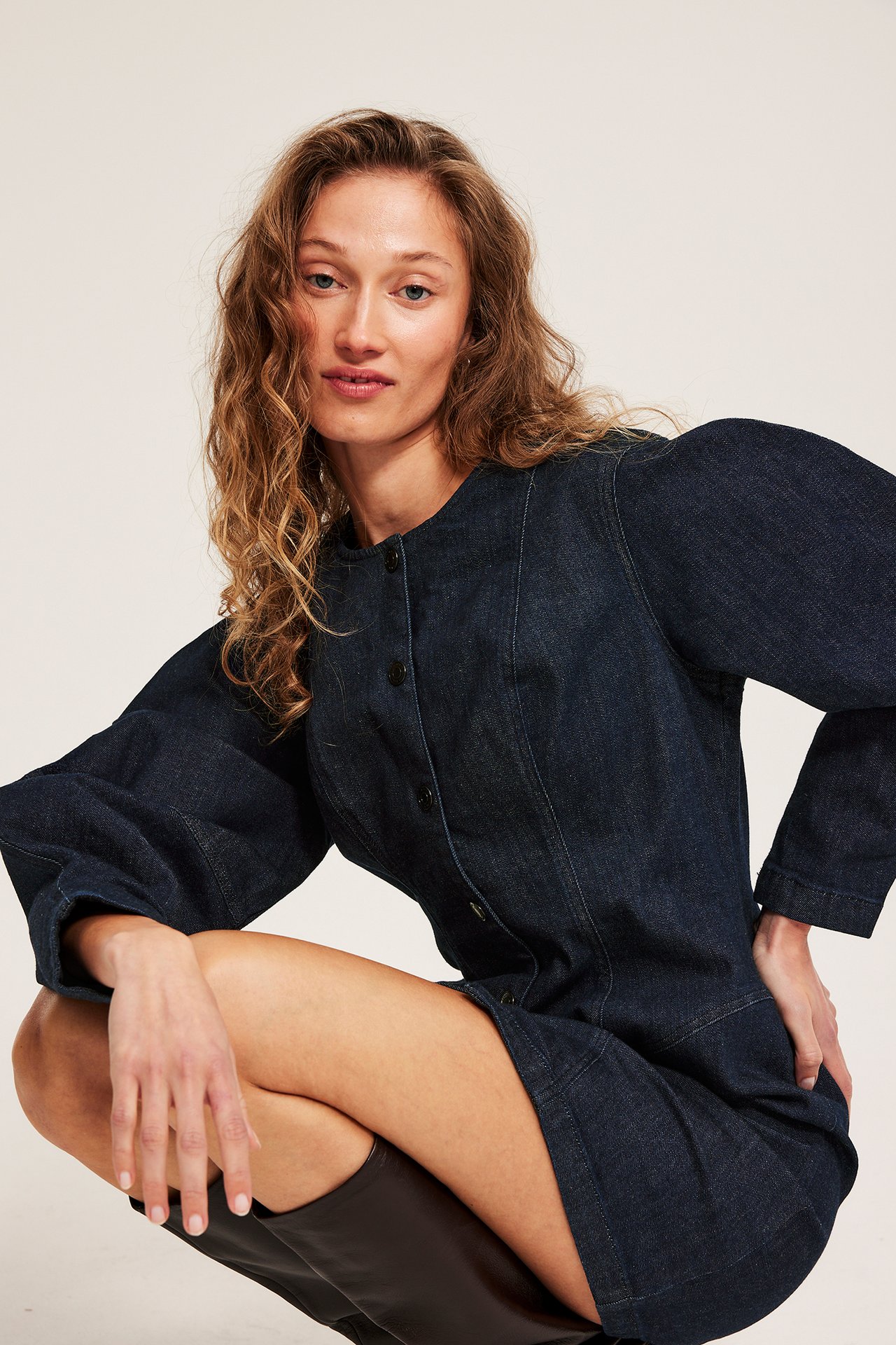 Frontvy, kvinna i mörk denim figurnära klänning med voluminösa ärmar, knappar och mörka stövlar.