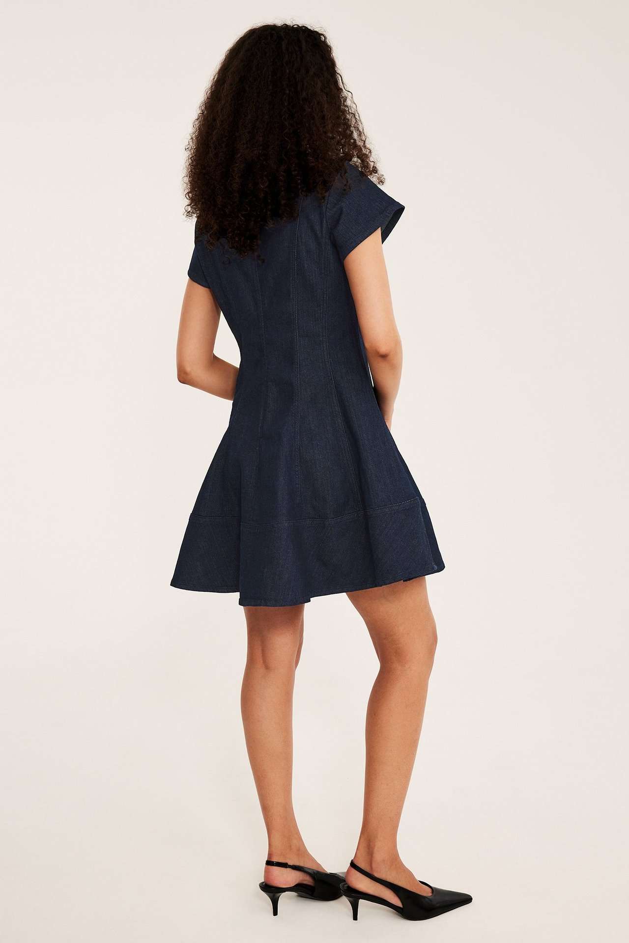 Naisen takakuva tummassa denim-A-linjaisessa mekossa saumoilla ja lyhyillä hihoilla, sekä mustissa slingback-korkokengissä.