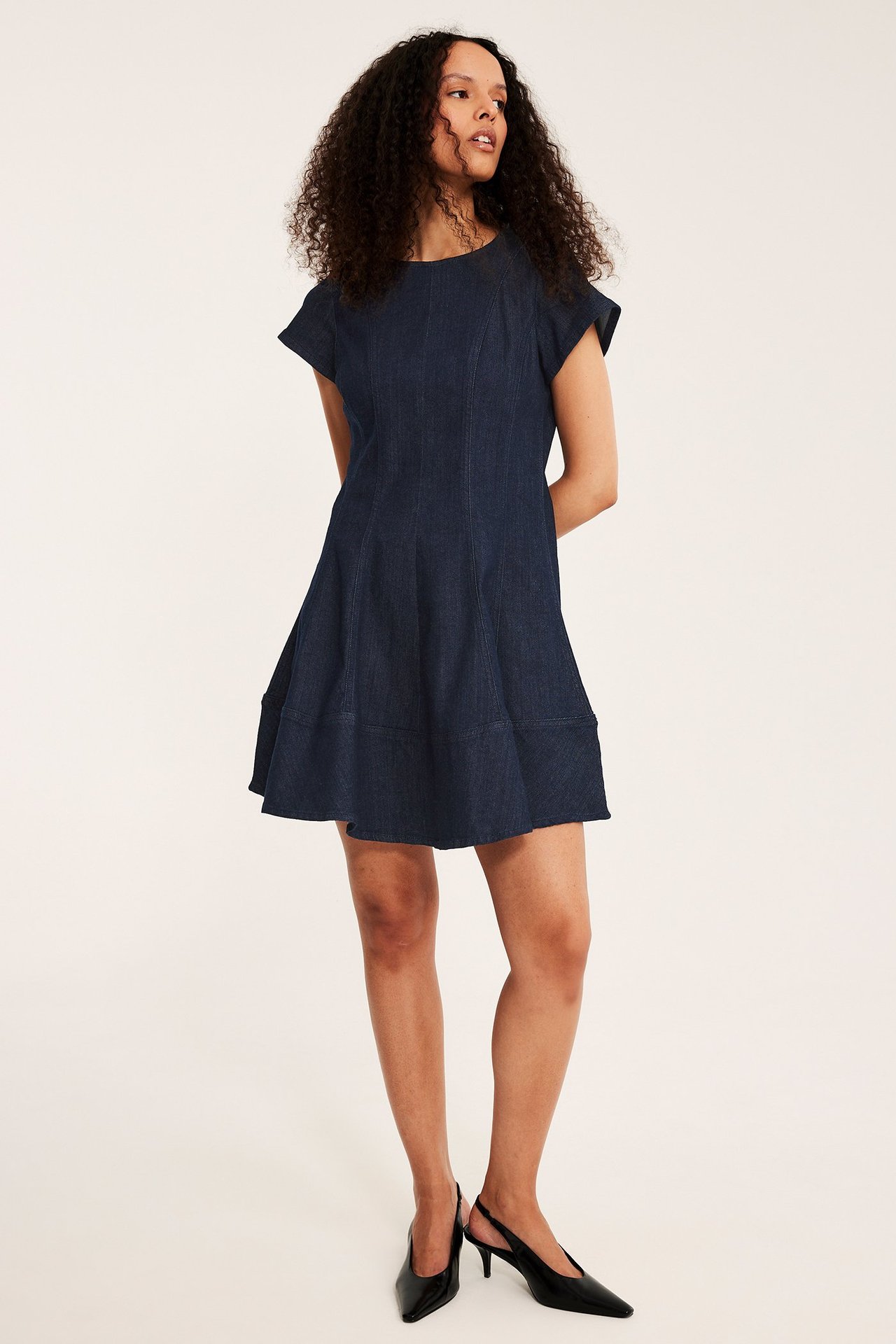 Edestä: Nainen yllään tummansininen A-linjainen farkkumekko saumoilla ja mustat avokkaat.