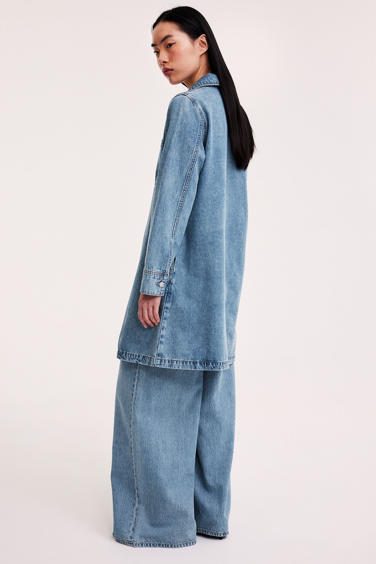 Sidebilde av en kvinne i en oversized lys blå dongerikjole stylet over vide dongeribukser.