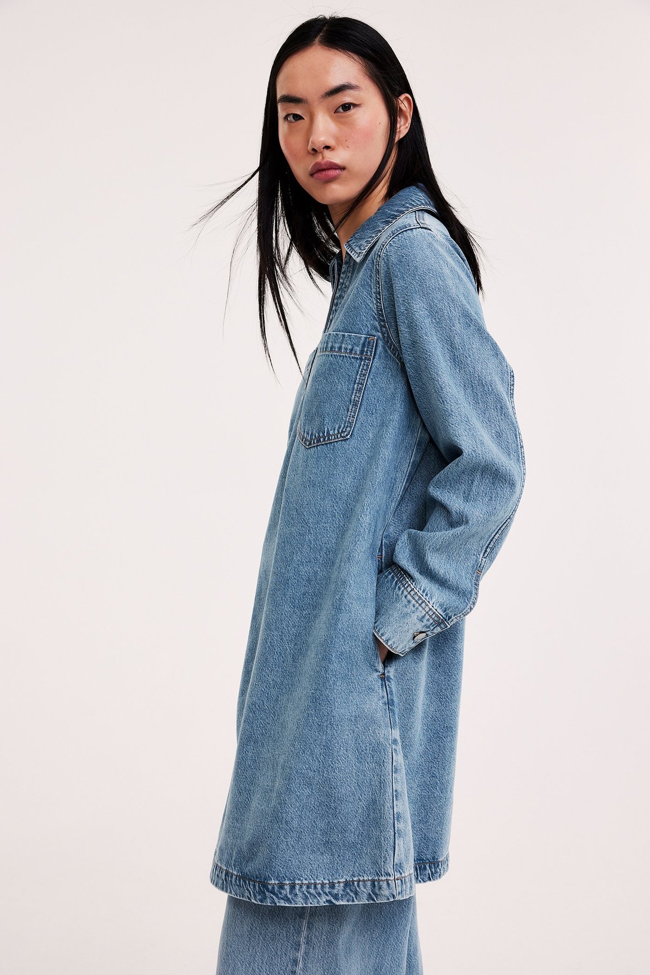 Sivukuva naisesta, yllään vaalea sininen farkkumekko kauluksella, rintataskuilla, ja farkkuhousut.