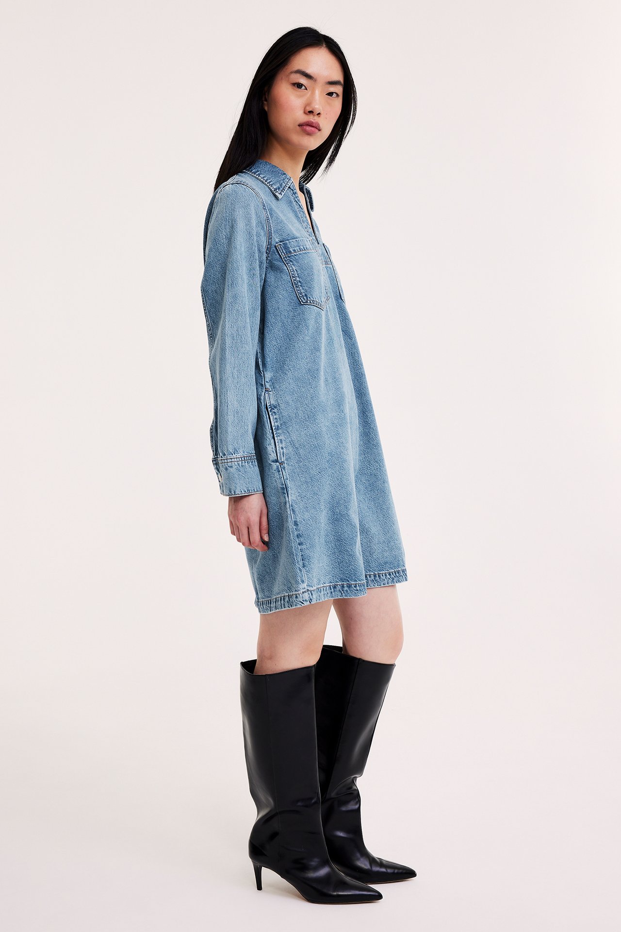 Sideprofil av kvinne i lys blå denimkjole med krage, brystlommer og svarte knestøvler.