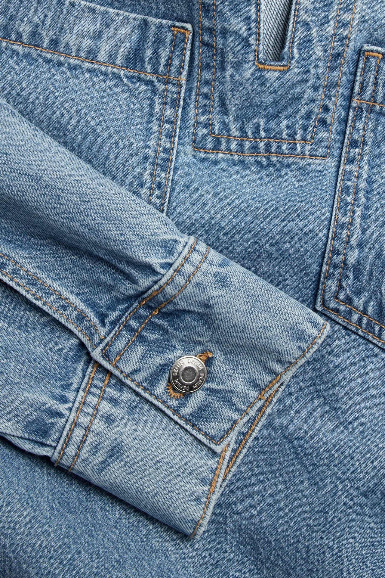 Lähikuva vaaleansinisestä farkkumekosta, jossa näkyy hihan mansetti hopeanapilla ja paikkataskut.