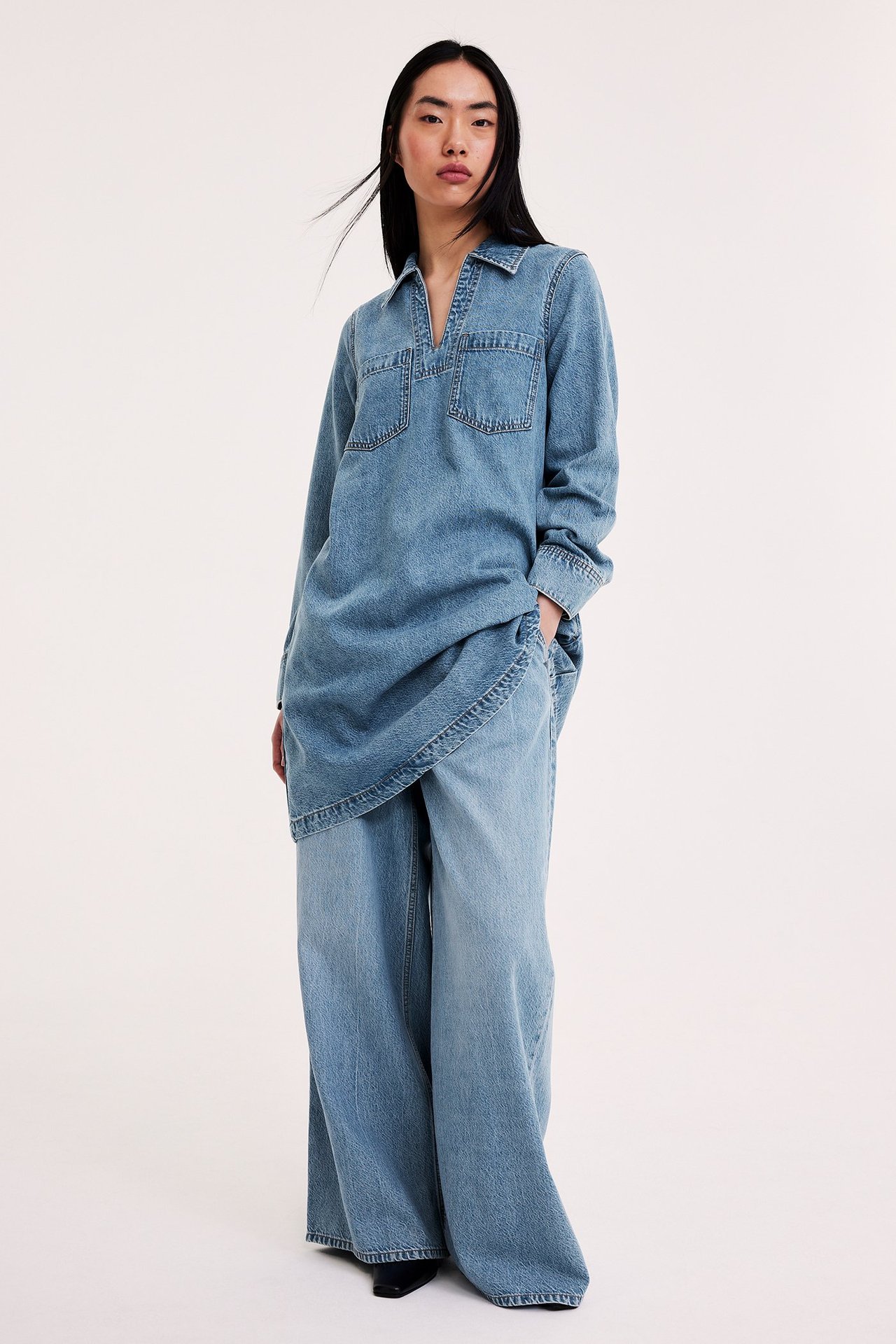 Nainen löysässä farkkutunikamekossa kauluksella ja rintataskuilla, leveät farkut alla.