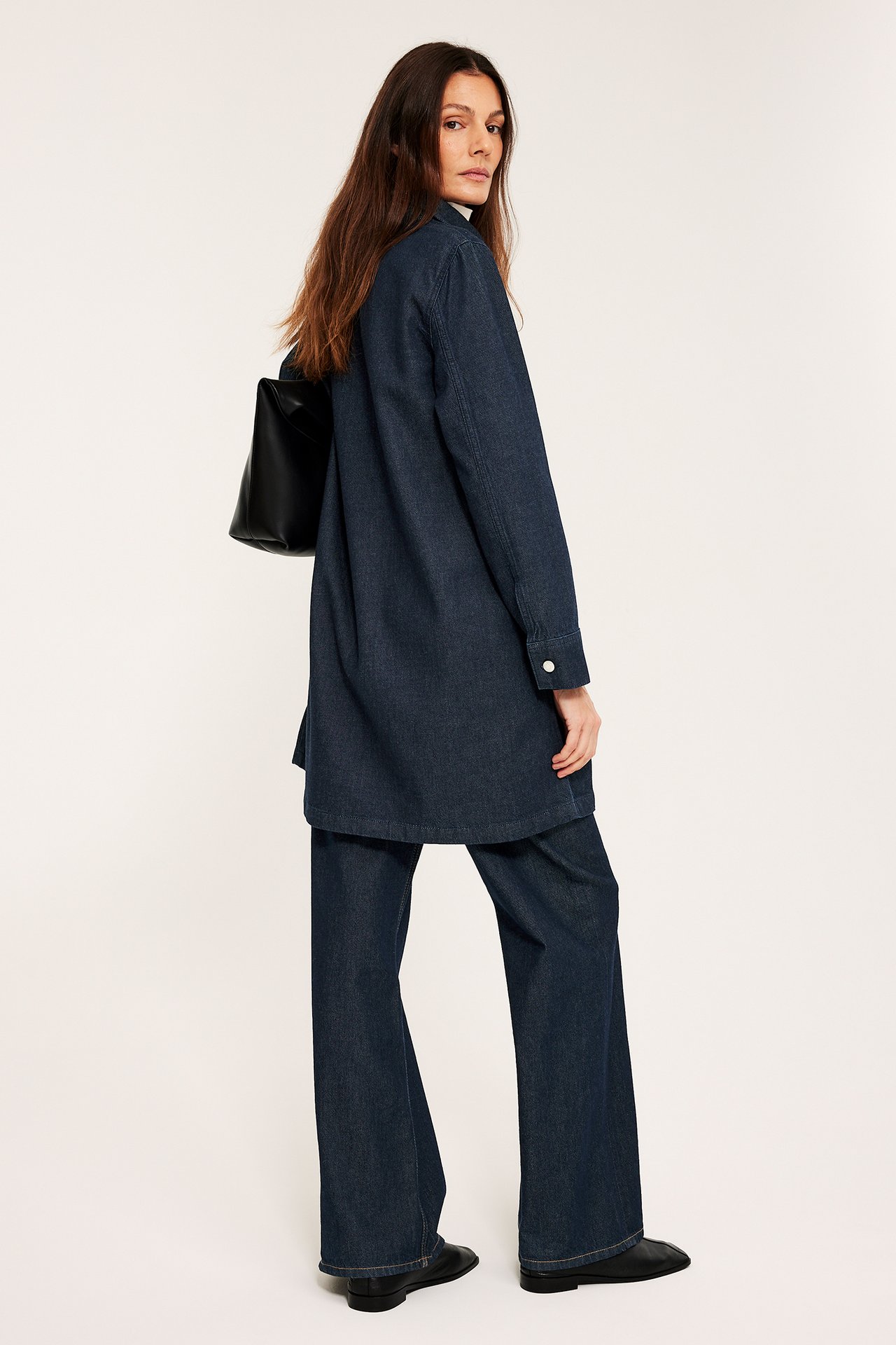 Kobieta, widok z boku, w ciemnej sukience jeansowej na pasujących spodniach, trzyma czarną torbę.