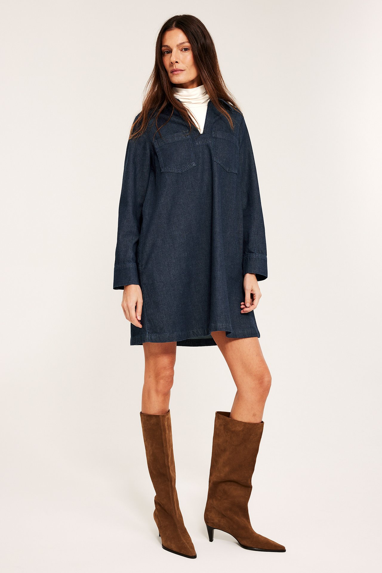 Widok z przodu kobiety w ciemnej jeansowej sukience mini z kieszeniami, białym golfie i brązowych kozakach.