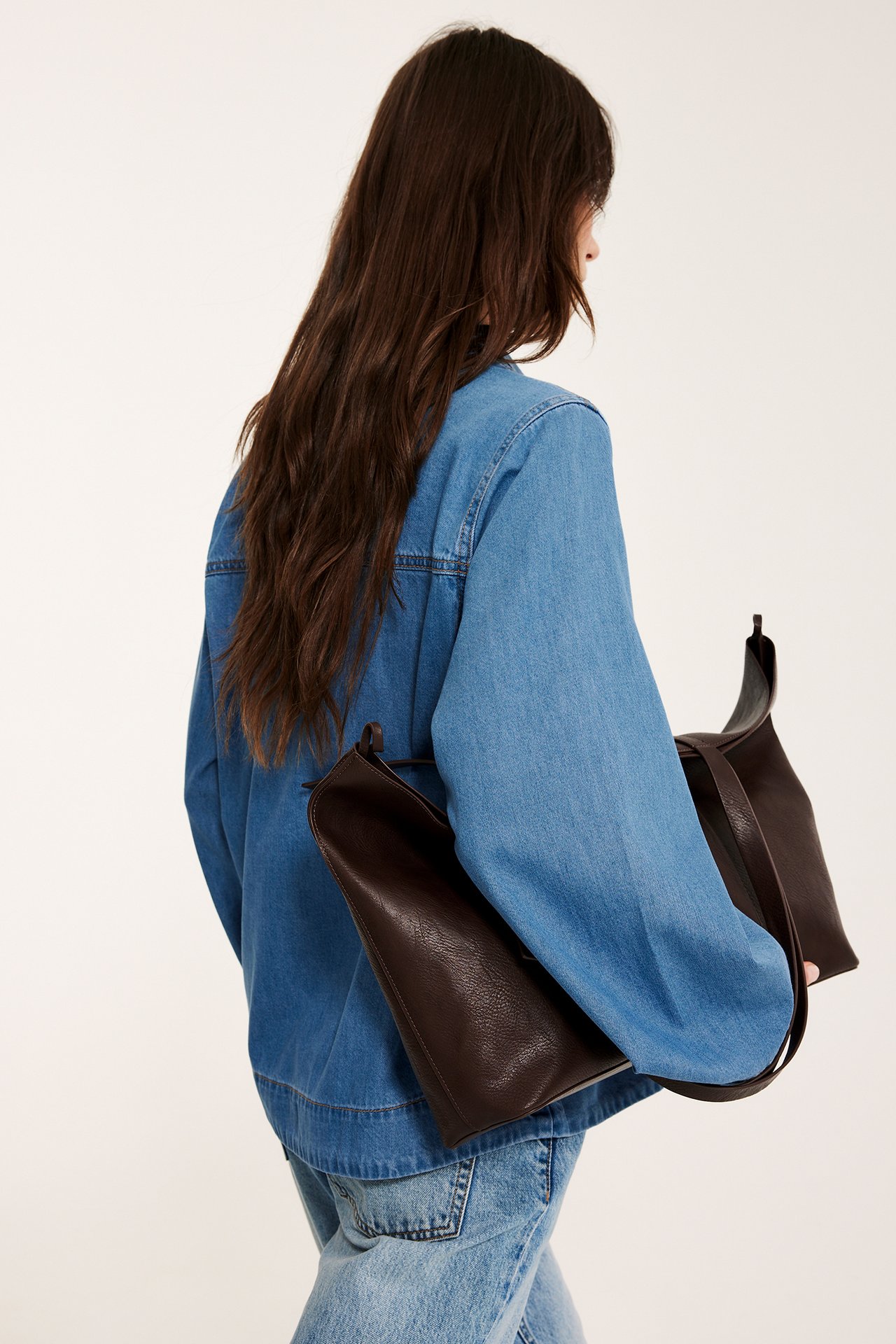 Nainen ylisuuressa sinisessä farkkupaidassa, vaaleansinisissä farkuissa, kantaen ruskeaa laukkua, takakuva.