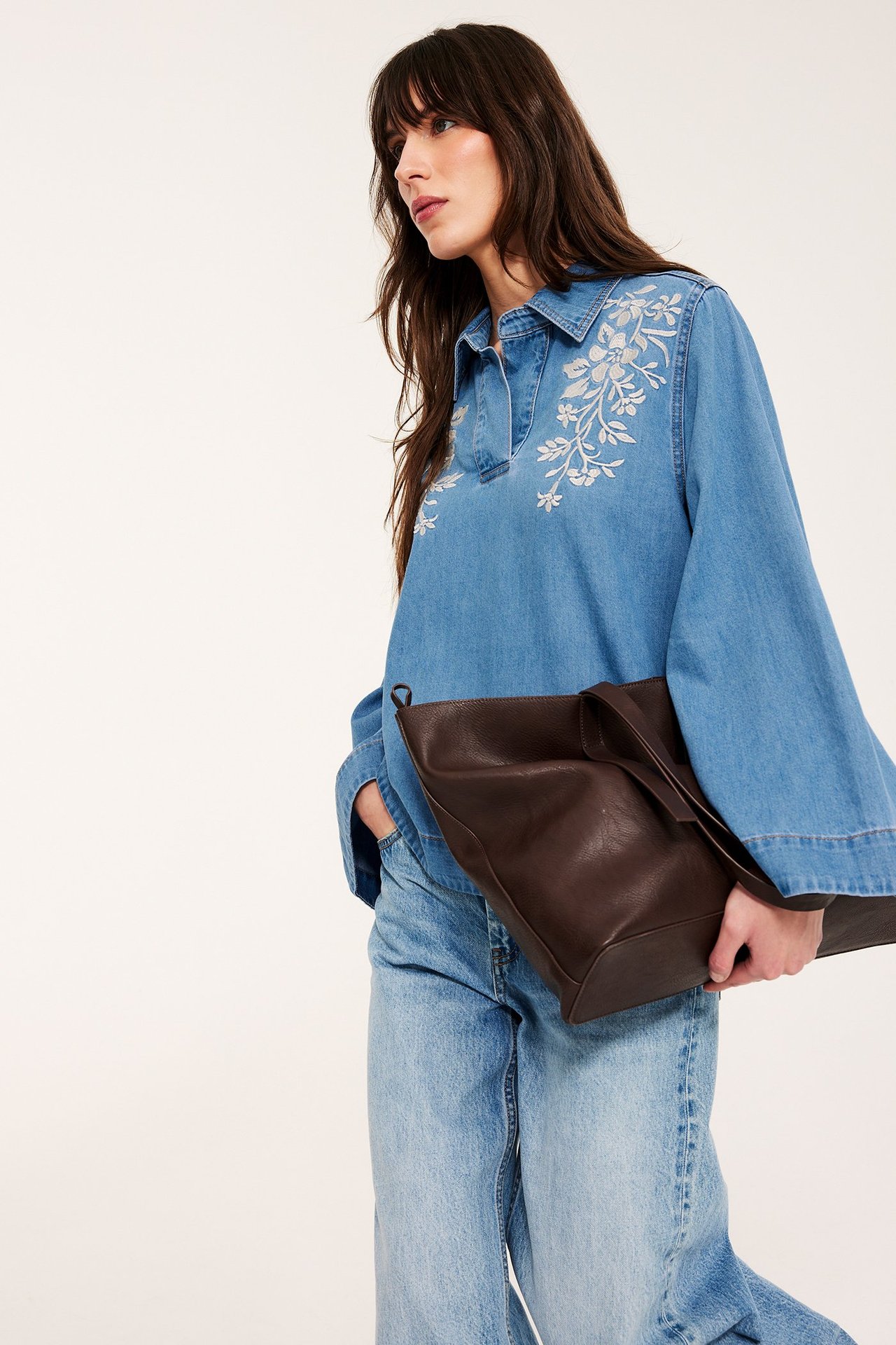 Nainen yllään brodeerattu vaaleansininen denim-paitapusero, vaaleat farkut ja pitää ruskeaa laukkua.