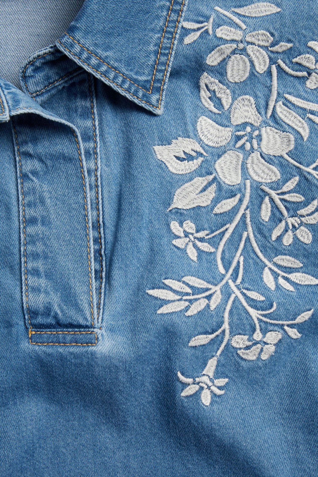 Zbliżenie denimowej koszuli popover z kołnierzem i białym kwiatowym haftem na piersi.