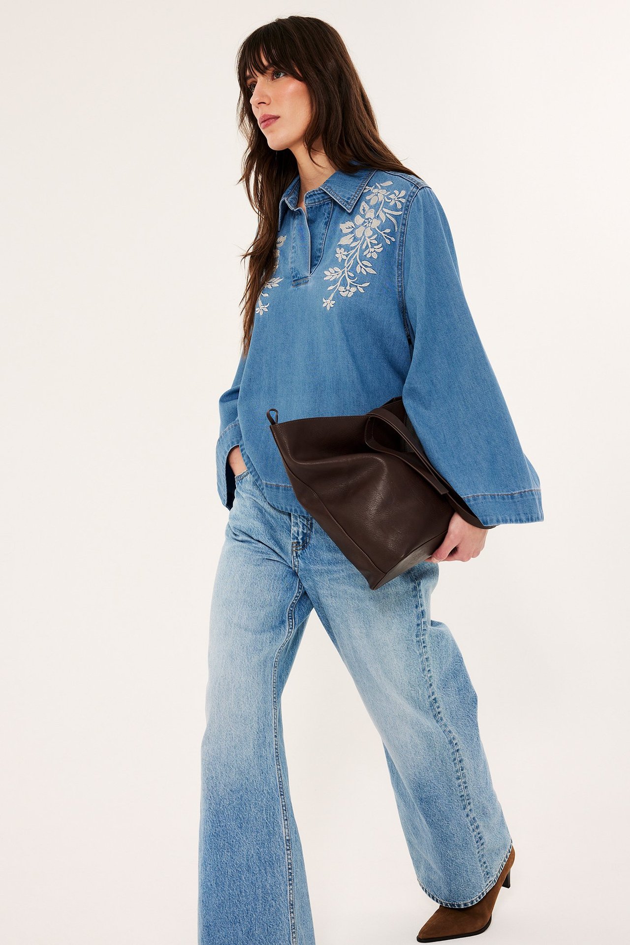 Edestä: Nainen sinisessä denim-puserossa, jossa valkoista kirjontaa, vaaleat farkut, pitää ruskeaa käsilaukkua.