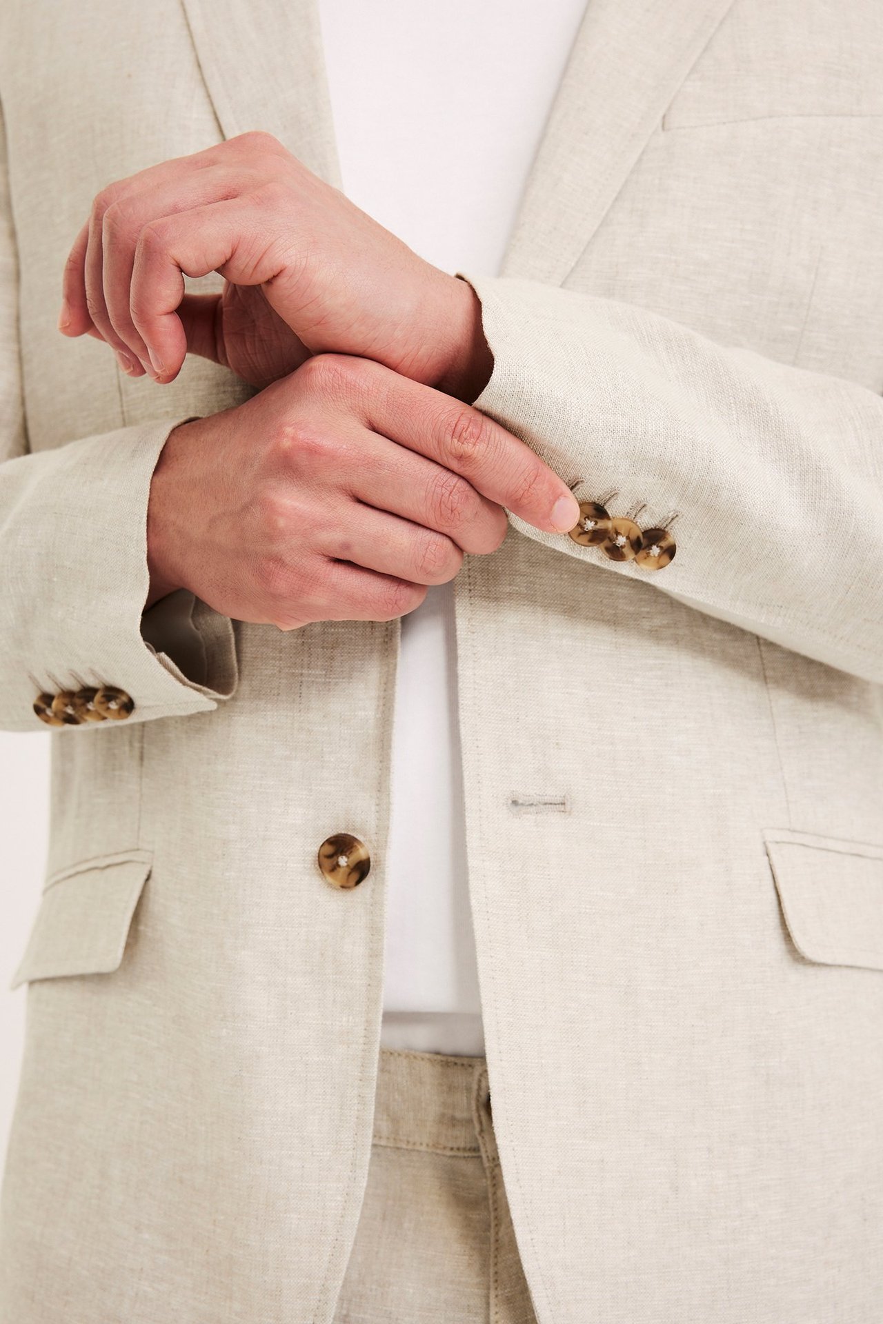 Nærbilde av mann som justerer sin beige regular fit linblazer, over hvit T-skjorte og matchende bukse.