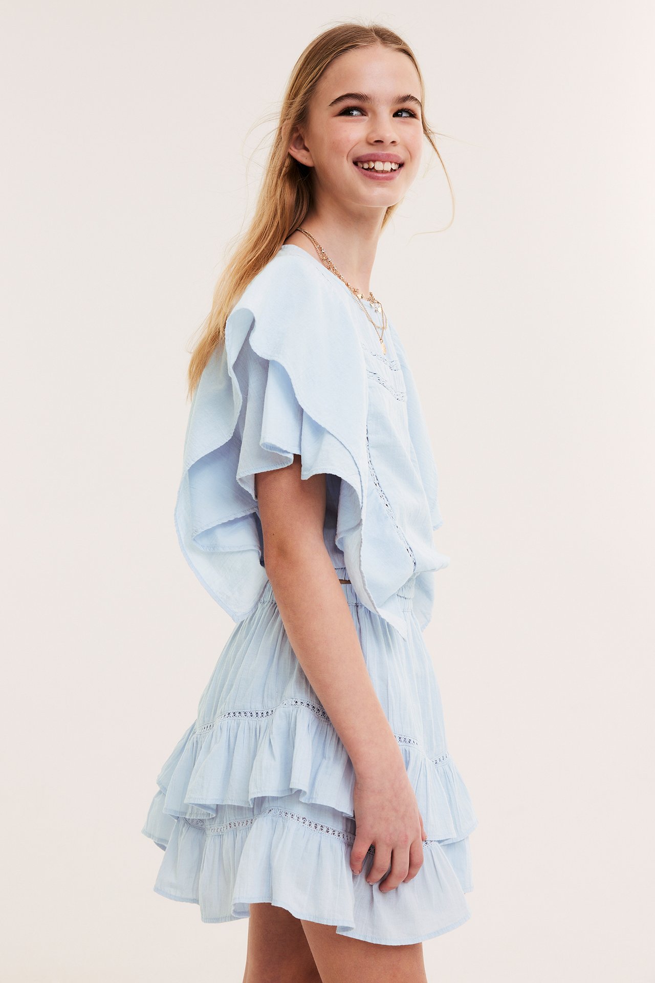 Sidovy av ett barn i en kort, blå volangkjol med spetsinsatser, matchande topp och guldhalsband.