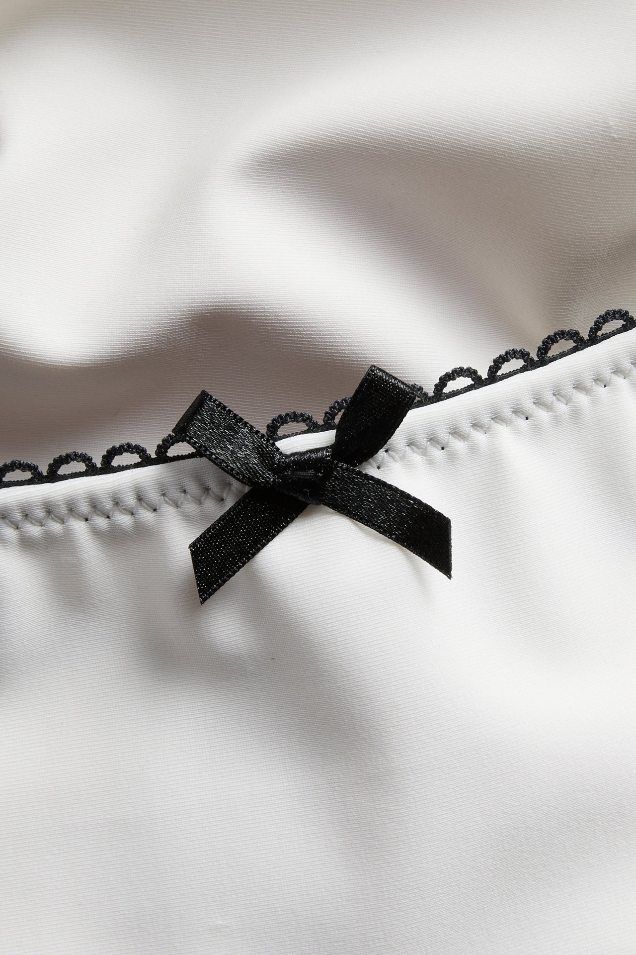 Zbliżenie na dół od bikini w kolorze off-white z czarną lamówką picot i małą czarną kokardką.