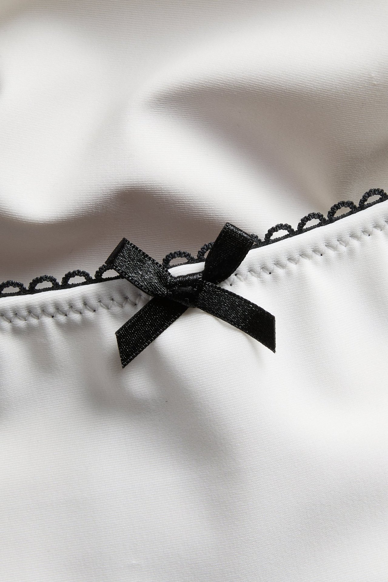Zbliżenie na dół od bikini w kolorze off-white z czarną lamówką picot i małą czarną kokardką.