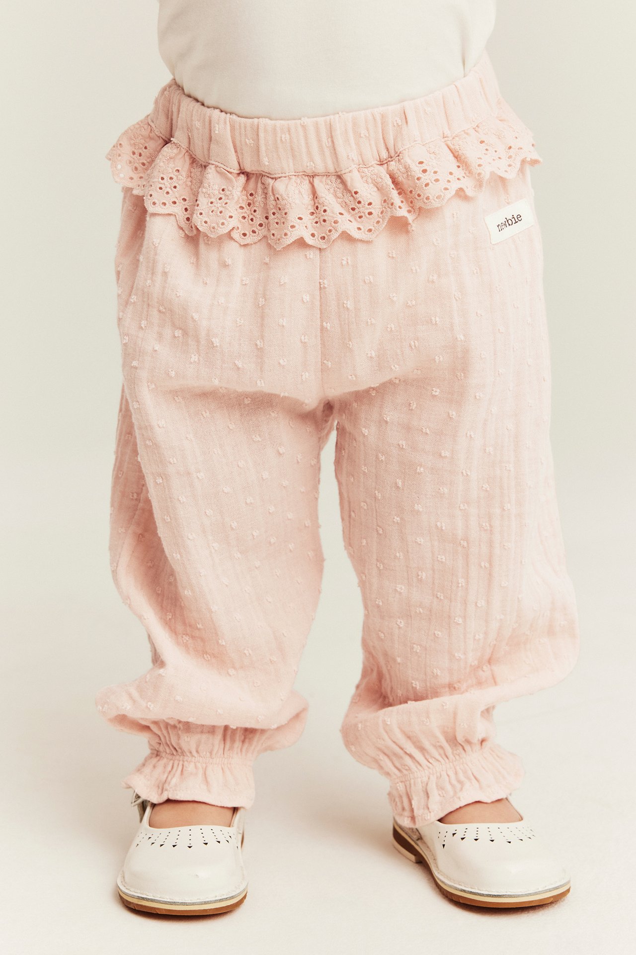 Framifrån: babys ben i ljusrosa strukturerade bomullsbyxor med spetsvolang i midjan och vita skor.