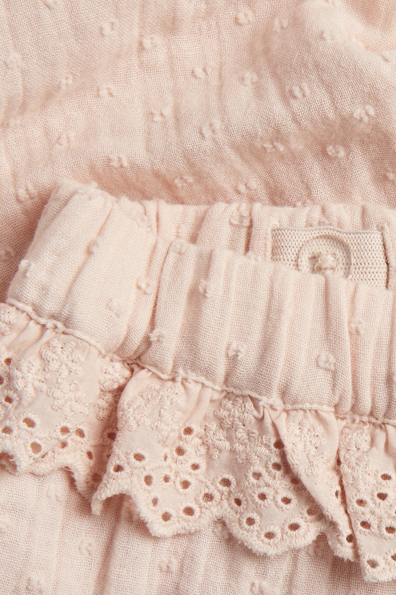 Nærbilde av lys rosa vevde bomullsbukser med prikkstruktur, elastisk midje og blondevolang.