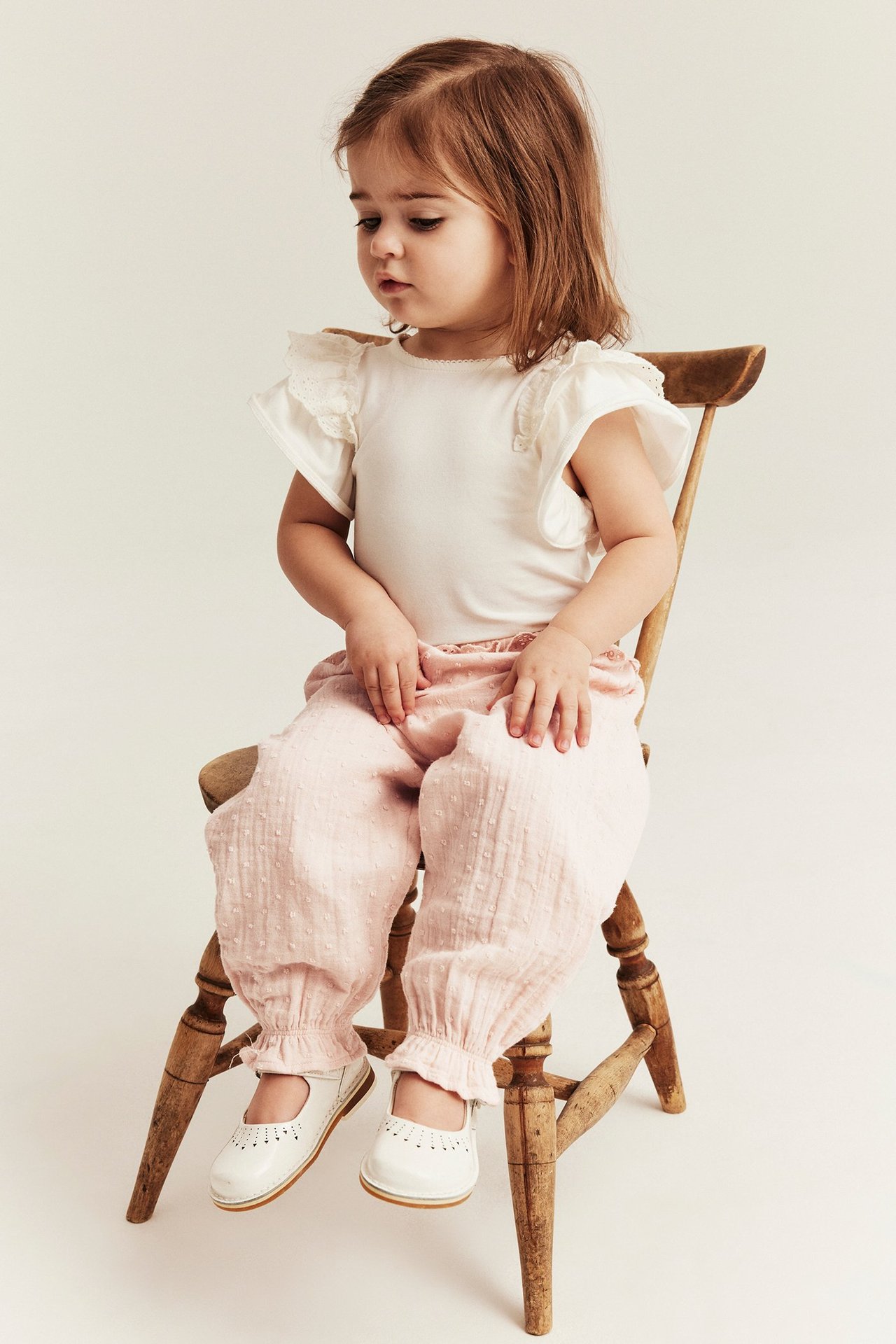 Framifrån: liten flicka i ljusrosa prickiga bomullsbyxor med volanger, vit topp och vita skor.
