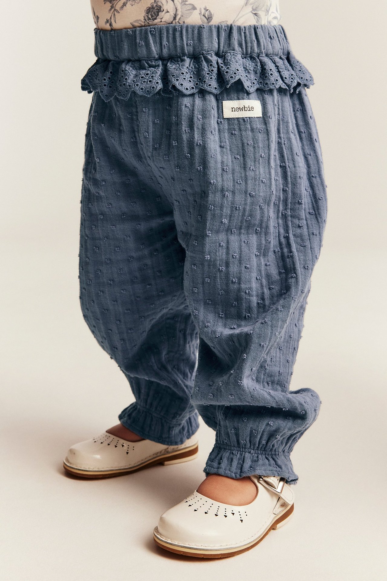 Nærbilde av baby i blå teksturerte bukser med blondedetaljer, elastisk midje og hvite sko.