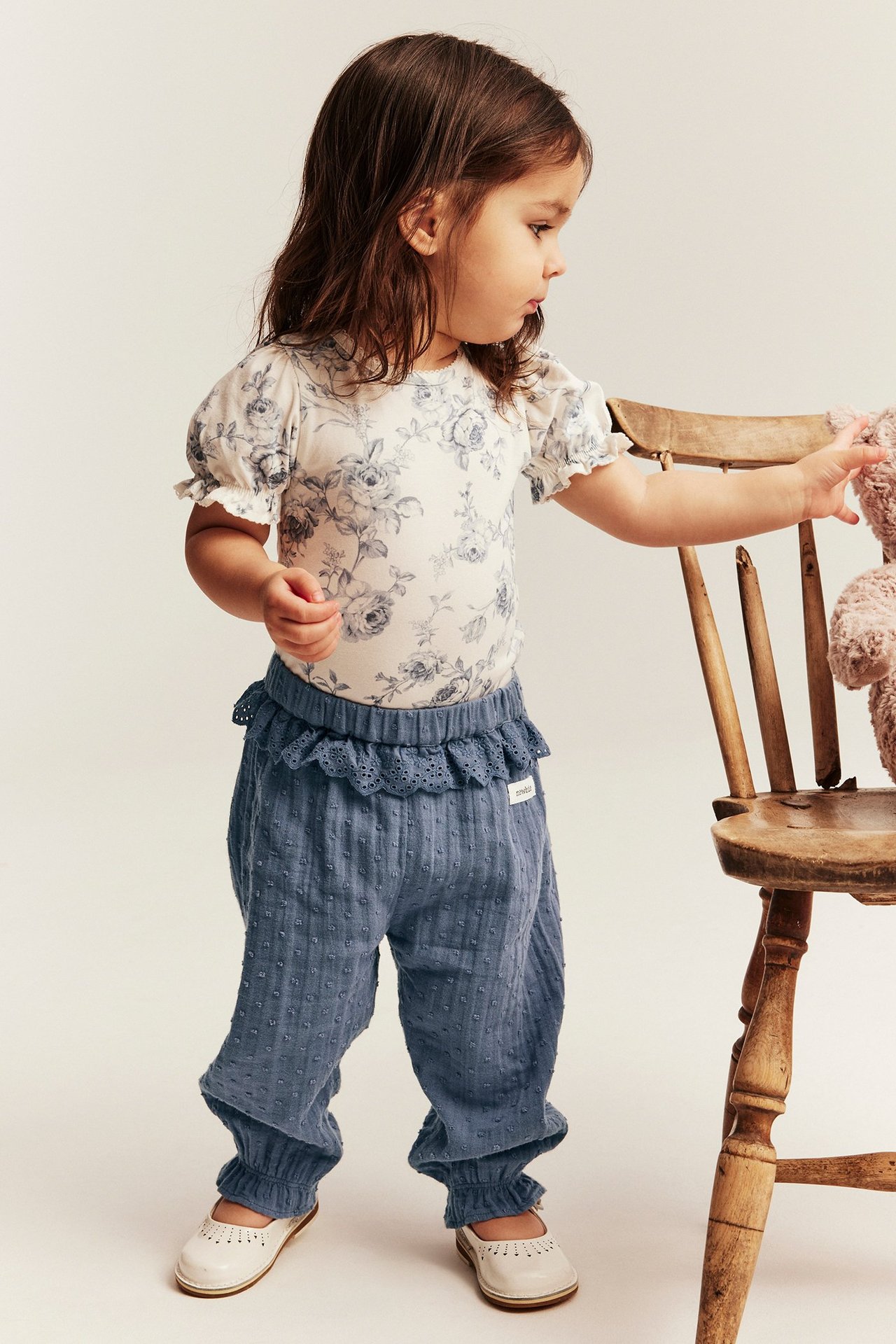 Forfra: barn i blå bomullsbukser med prikkstruktur og blonder, blomstret topp, hvite sko.
