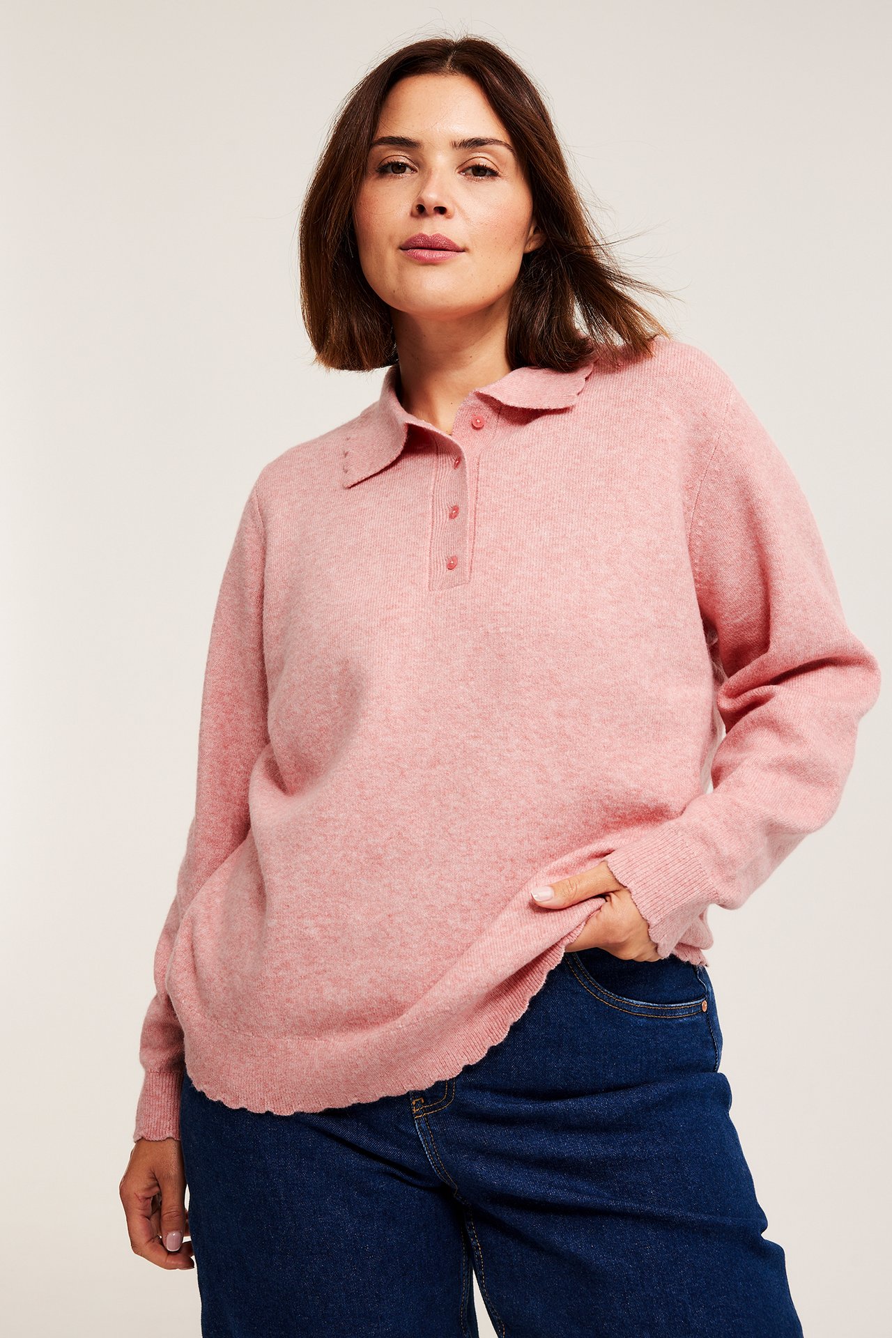 Framifrån, kvinna i rosa melerad finstickad tröja med vågiga kanter och mörkblå jeans.