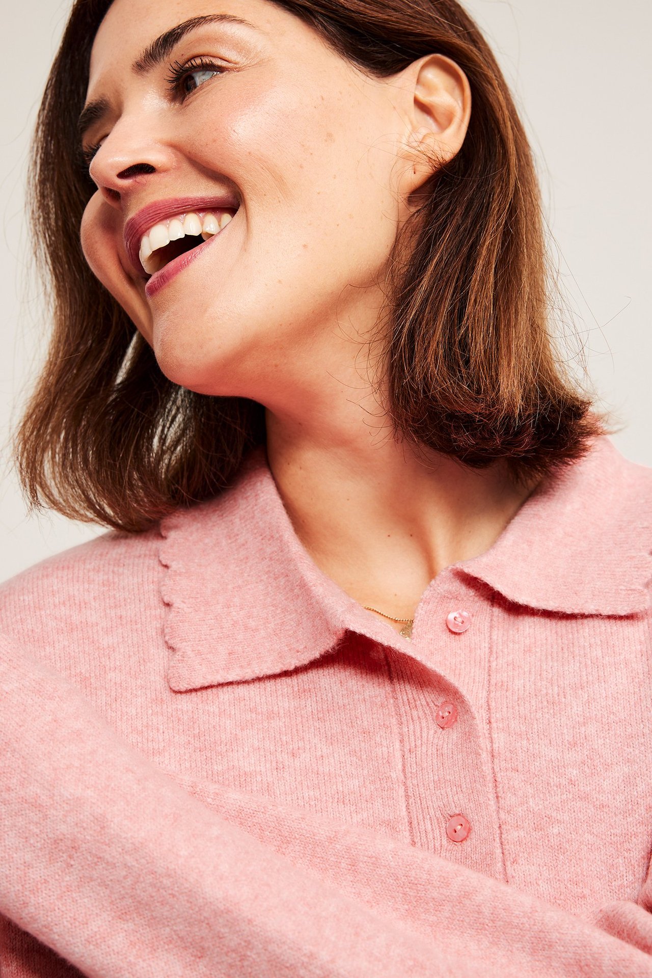 Närbild av en kvinna i rosa finstickad tröja med knappslå och vågig krage, med en tunn guldkedja.