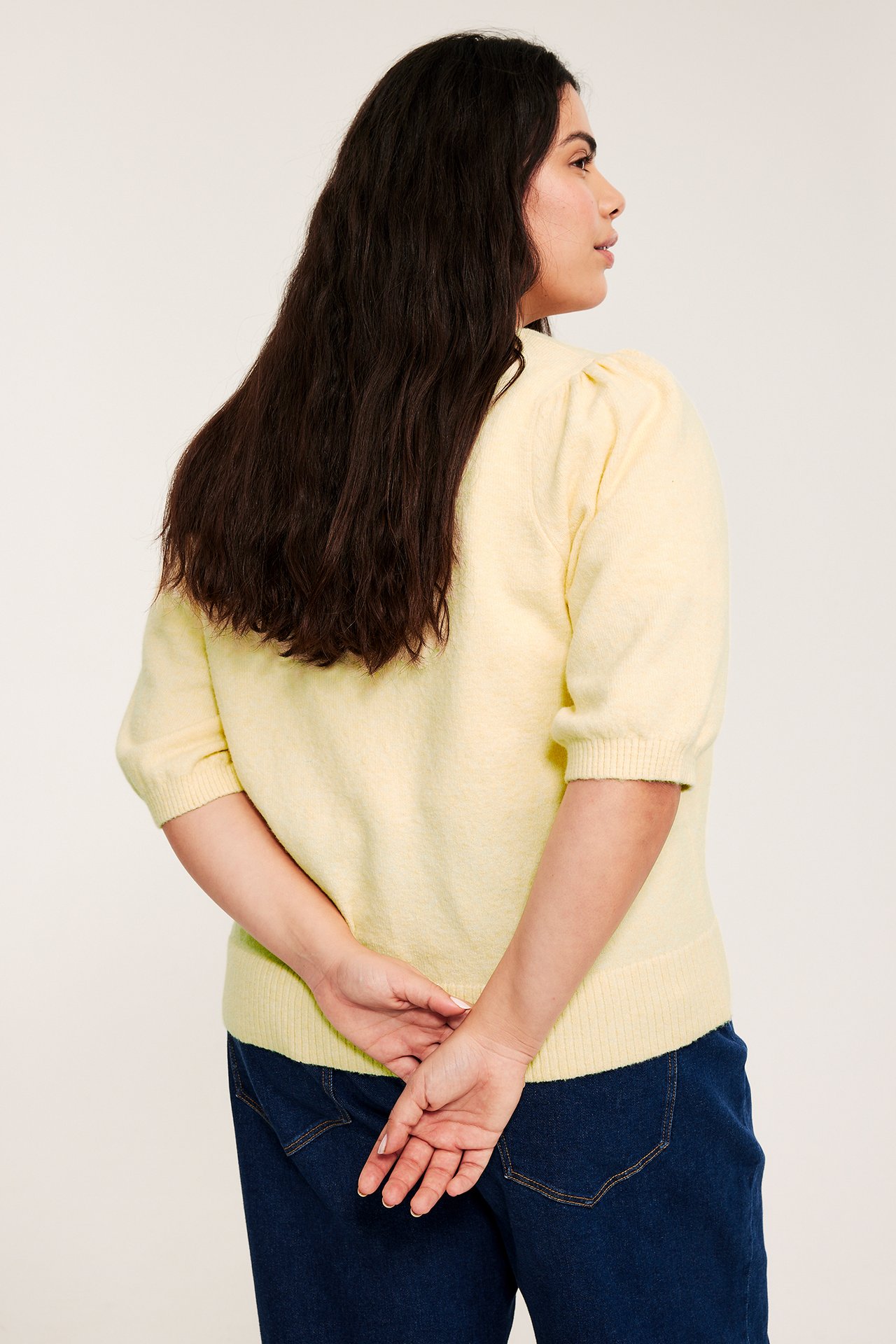 Widok z boku/tyłu: kobieta w jasnożółtym melanżowym kardiganie z krótkimi bufiastymi rękawami, ciemnoniebieskie jeansy.