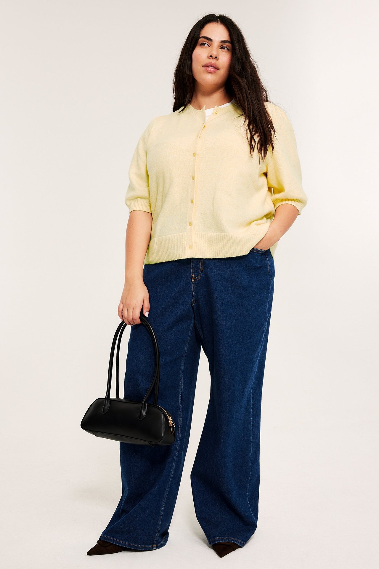 Nainen vaaleankeltaisessa pörröhihaisessa neuletakissa, tummansinisissä farkuissa, pitää mustaa laukkua.