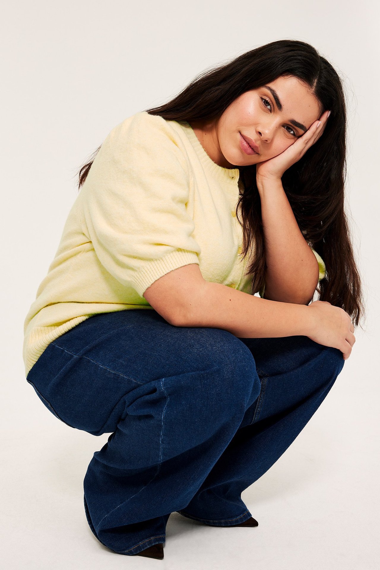 Nainen yllään vaaleankeltainen meleerattu lyhythihainen neuletakki ja tummansiniset leveälahkeiset farkut.