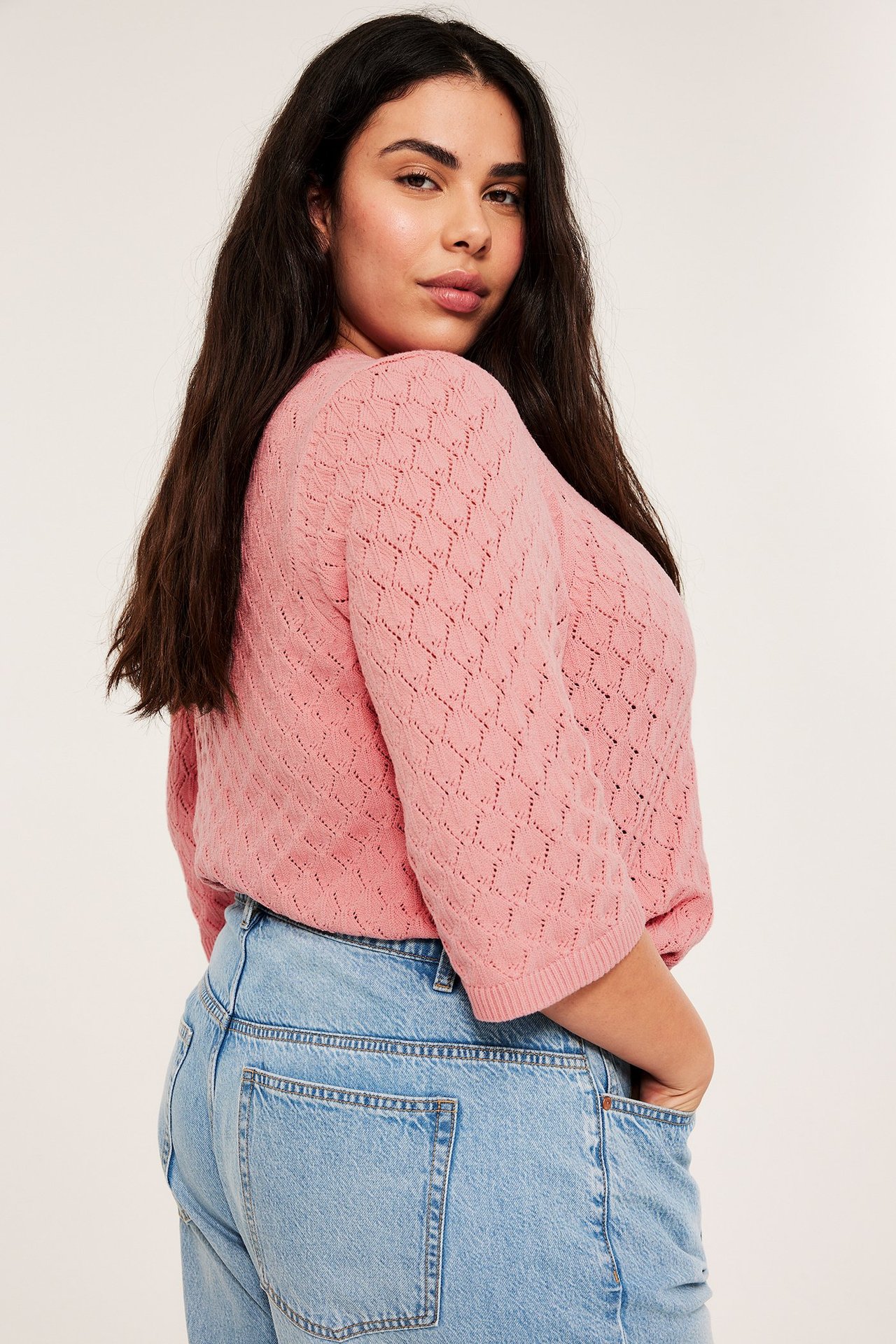 Sidovy av en kvinna i rosa kortärmad spetsstickad tröja och ljusblå jeans.