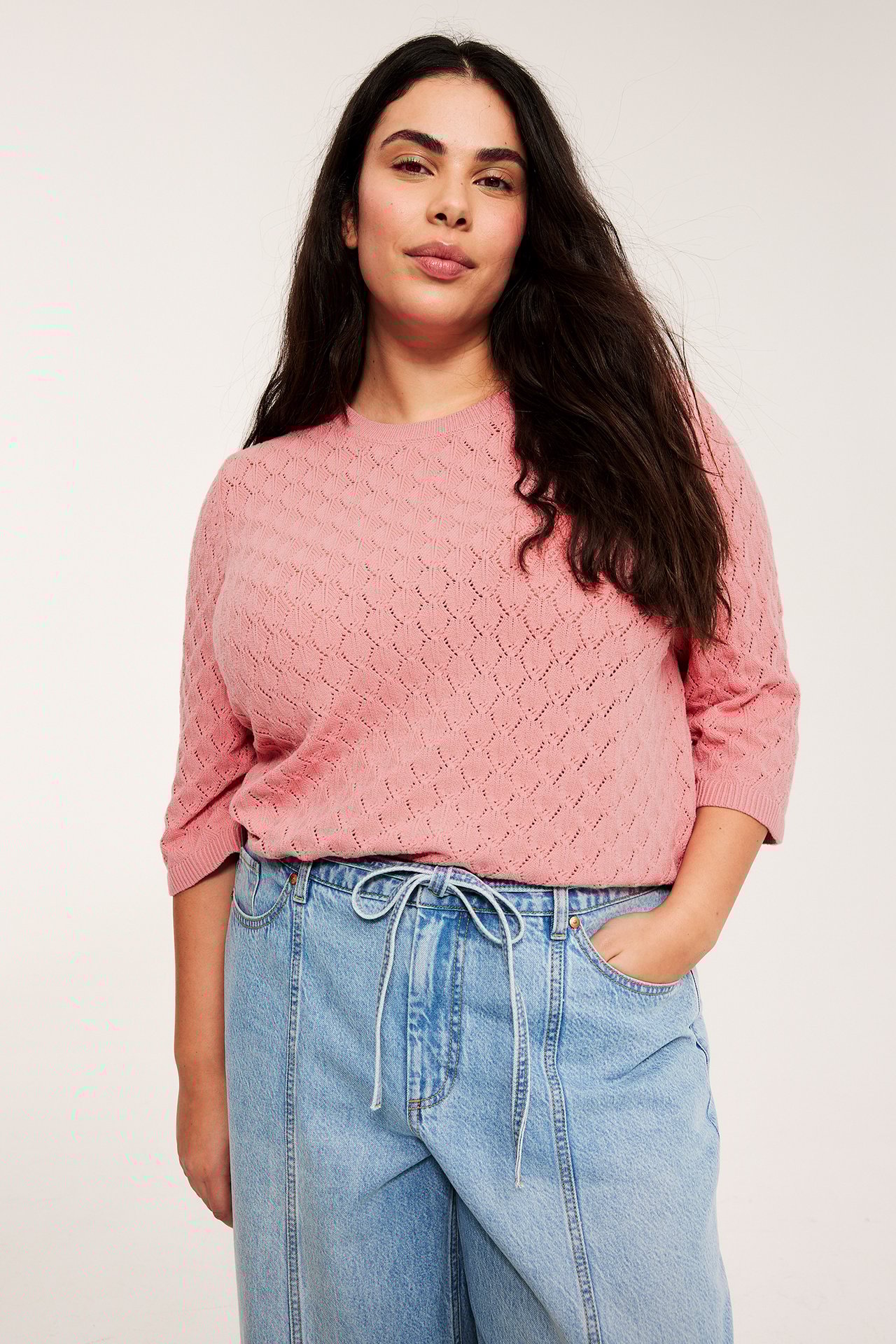 Nainen yllään vaaleanpunainen pitsineulottu lyhythihainen neulepaita ja vaaleansiniset kiristysnyörilliset farkut.