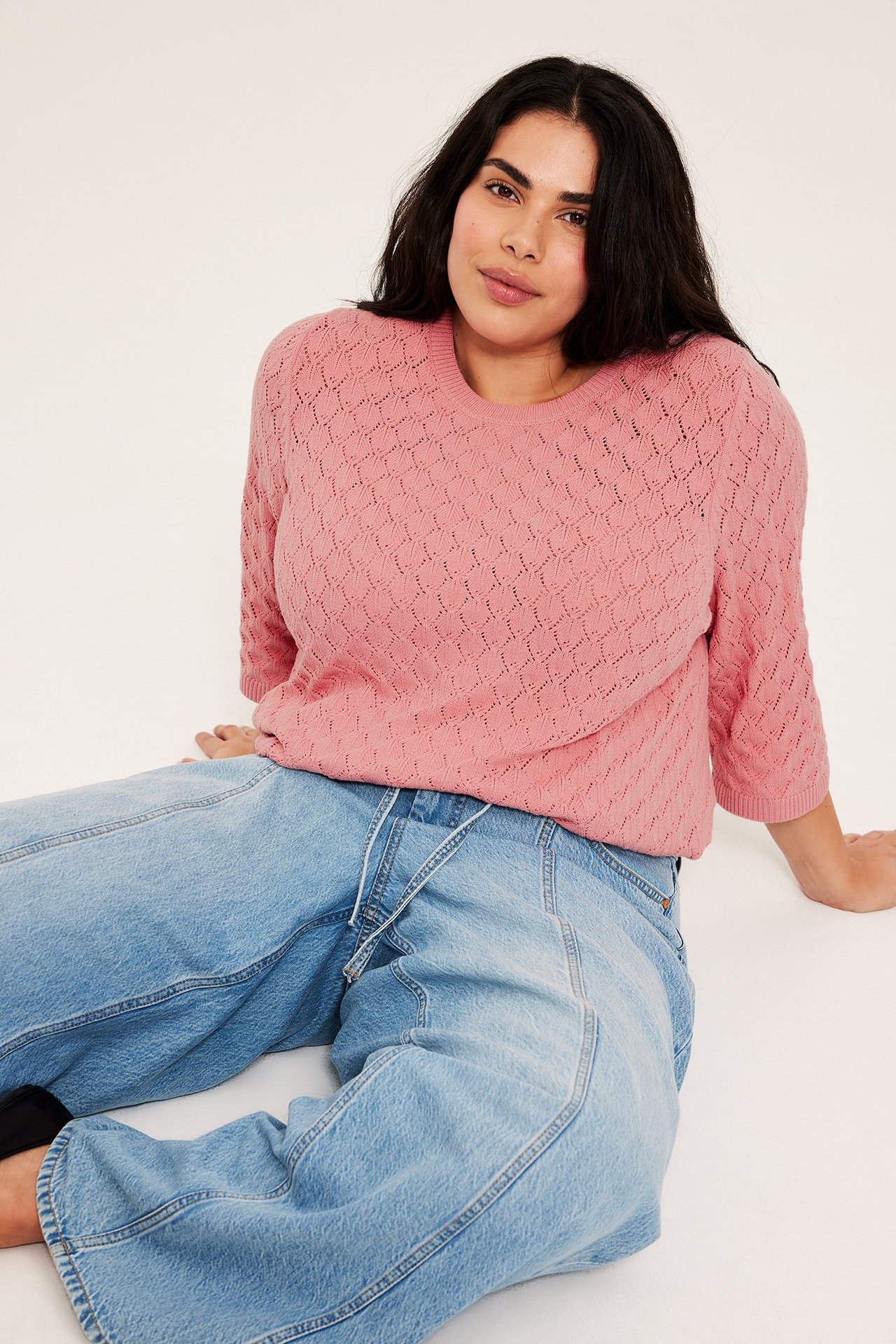 Widok z przodu kobiety w różowym ażurowym swetrze z krótkim rękawem i jasnoniebieskich jeansach wide-leg.