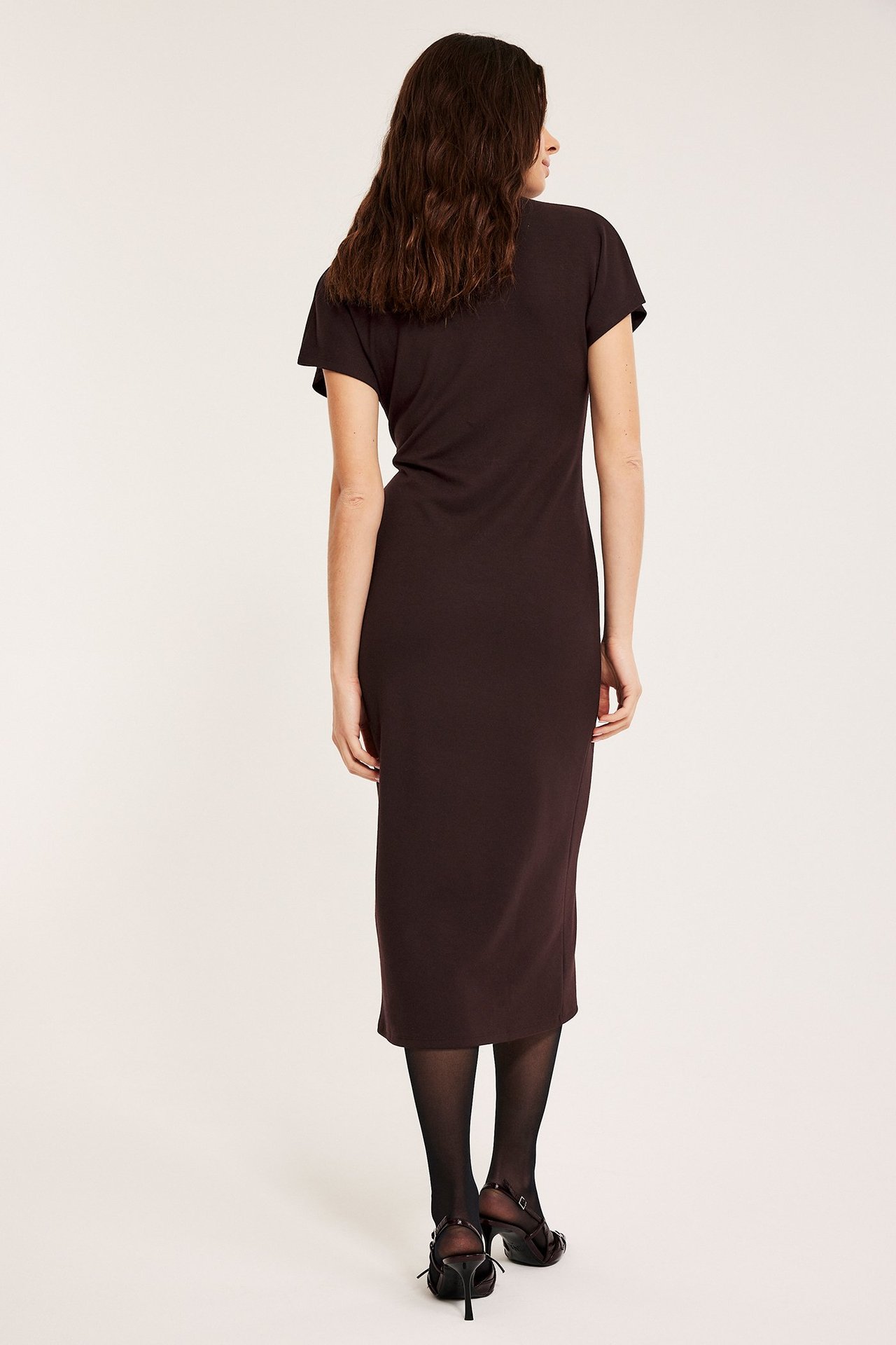 Takaapäin: nainen viininpunaisessa jersey-pitkämekossa, lyhyillä hihoilla, mustissa sukkahousuissa ja korkokengissä.