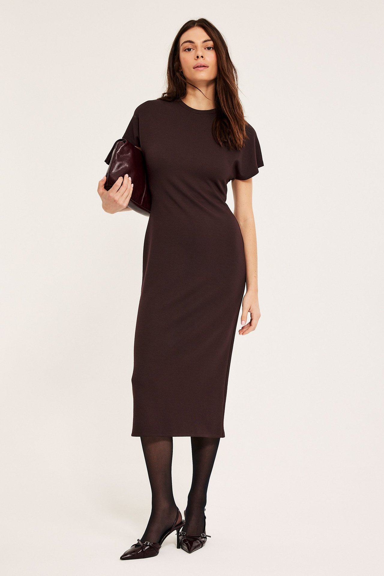 Edestä: Nainen yllään viininpunainen trikoo-midimekko, pidellen yhteensopivaa clutchia, mustat sukkahousut ja korot.