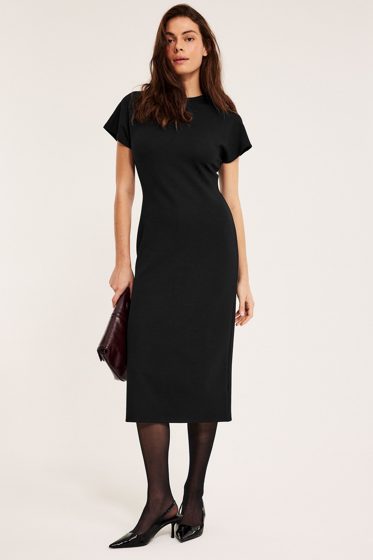 Widok z przodu kobiety w czarnej sukience dżersejowej, ciemnoczerwona kopertówka, czarne rajstopy, szpilki.