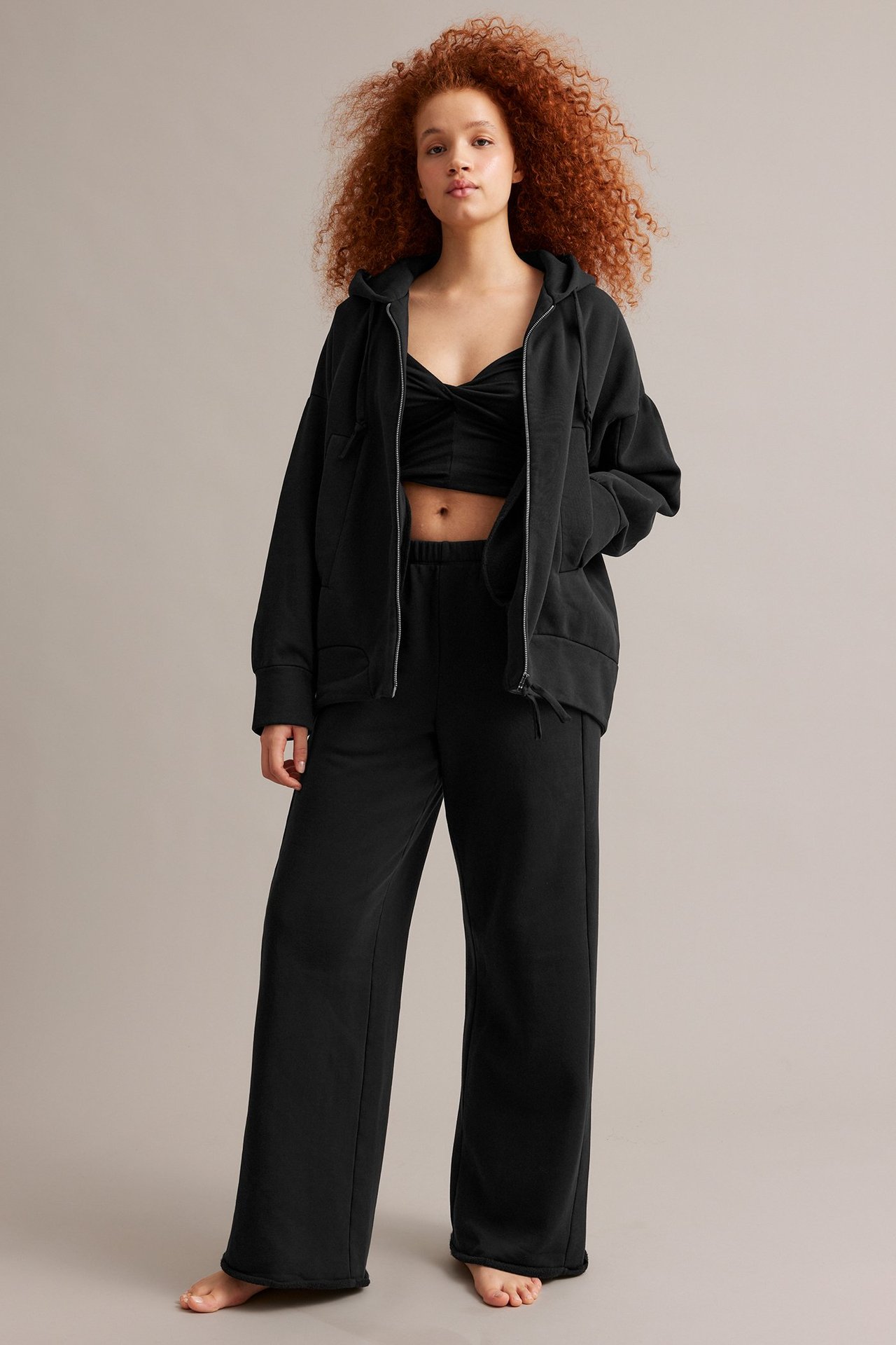 Forfra: kvinne i åpen svart oversized hettegenser, svart crop top og svarte vide joggebukser.