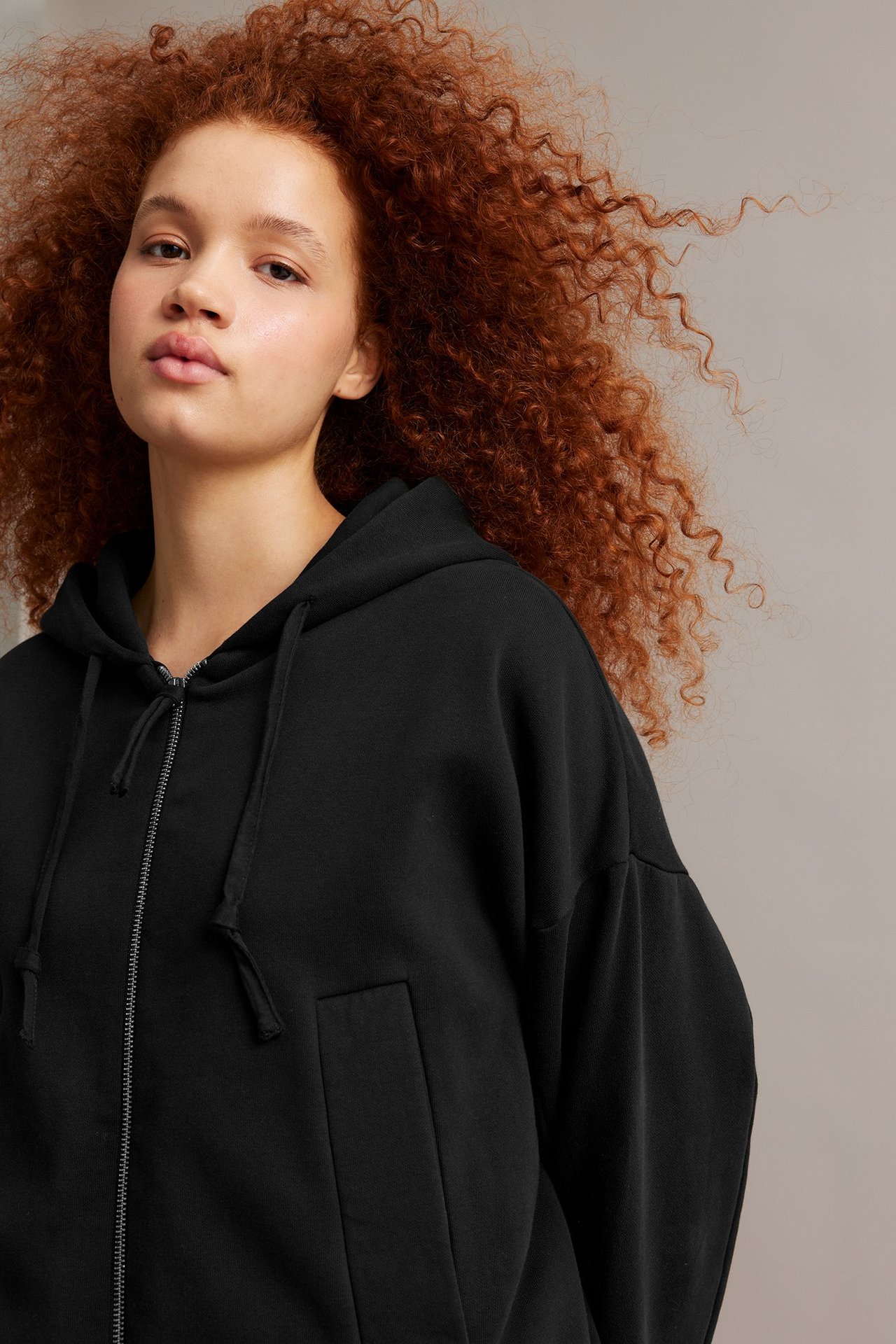 Nærbilde forfra: Kvinne med svart oversized hettegenser med glidelås og frontlommer.