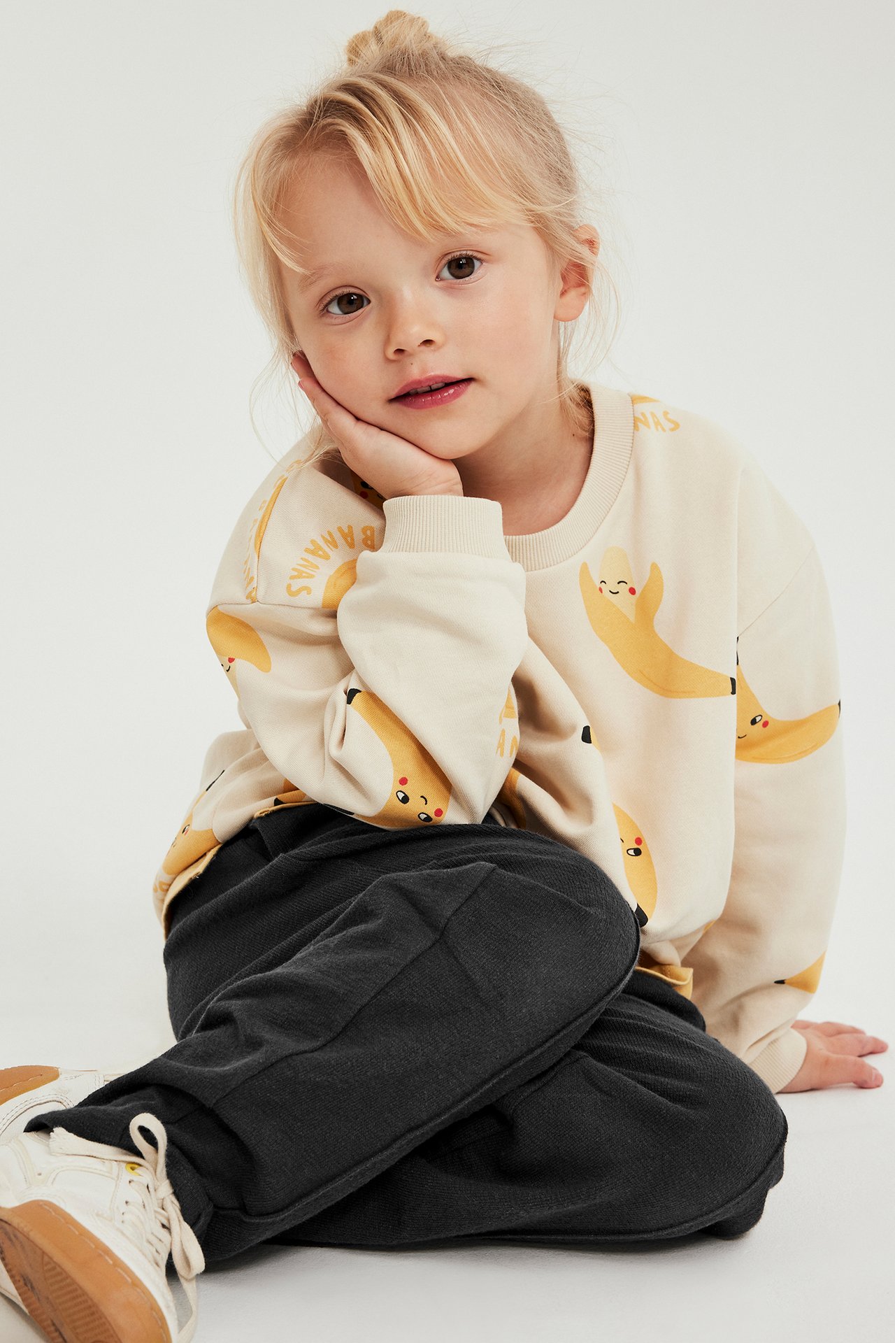 Framifrån: Ett barn i svarta texturerade mjukisbyxor, gräddfärgad bananmönstrad sweatshirt och vita sneakers.